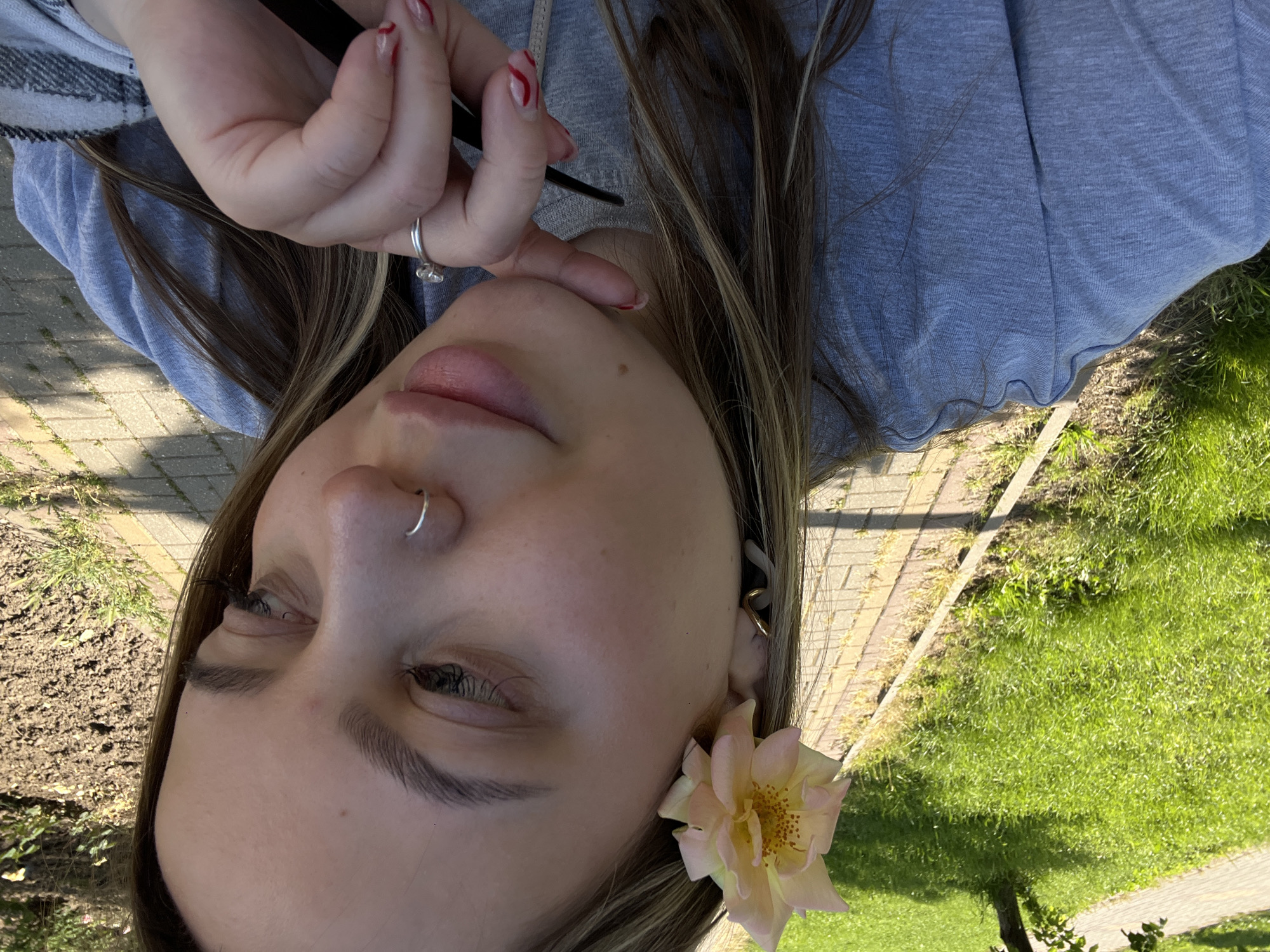 The image depicts a close-up outdoor portrait of a young adult female, confirmed to be in Novopokrovskaya, Russia. The subject is positioned facing slightly to the right of the frame, with her gaze directed downwards and to the right. She has light brown hair with blonde highlights, visible across her shoulders and on the right side of her face. A single, light pink rose with a yellow center is tucked behind her left ear. Her left ear is adorned with a gold-colored, abstract-shaped earring. A silver-colored hoop nose ring is present in her right nostril. Her fingernails on her visible right hand display a red and white striped or swirled pattern. She wears a light blue, possibly heathered, long-sleeved top, appearing to be a sweatshirt or casual knit. A grey and white plaid pattern is visible on the cuff or sleeve of a garment on her right arm. Her right hand is raised, with her index finger resting near her chin in a contemplative or relaxed gesture. A silver-colored ring with a clear stone is on the ring finger of her right hand. She holds a slim, dark-colored object, resembling a pen or stylus, between her right thumb and index finger.
The background shows an outdoor setting on a sunny day. To the left, there is a manicured green lawn, brightly lit by direct sunlight, bordered by a paved pedestrian path made of rectangular light-colored bricks. A raised concrete curb separates the grass from the path. Further to the left, a small tree trunk and dappled shadows are visible on the grass. To the right, beyond the path, an area of disturbed or tilled earth is visible, with sparse green vegetation.