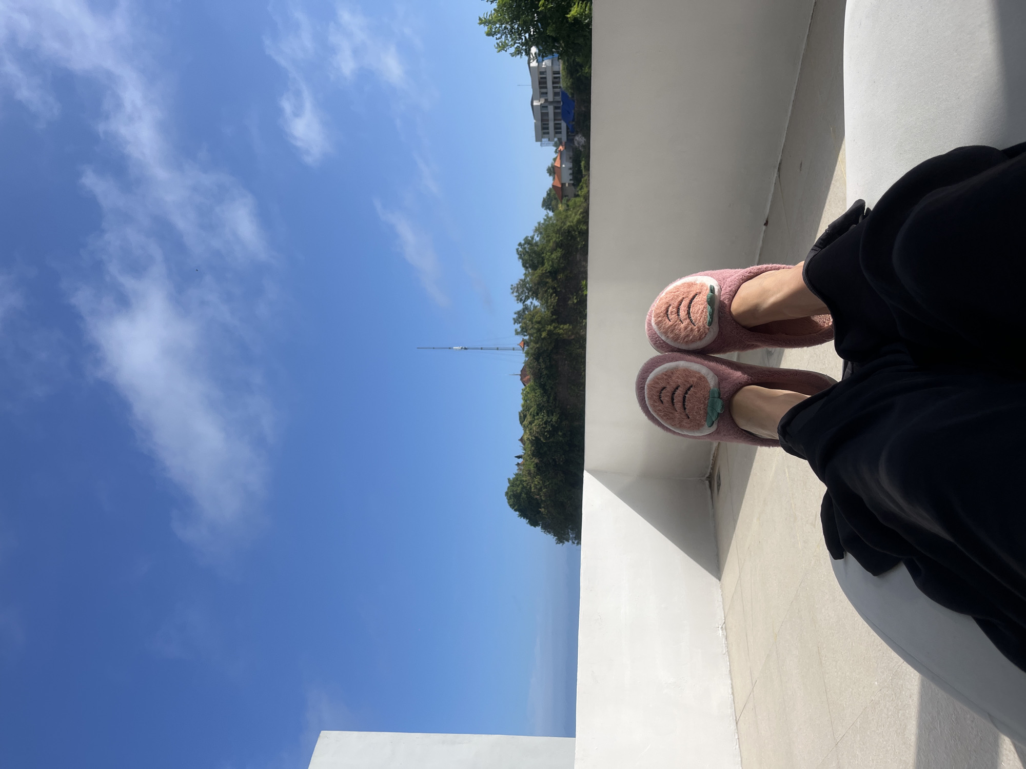 A point-of-view shot shows a person sitting on a white tiled surface, likely a balcony or patio, overlooking an outdoor scene. The person is wearing black pants and pink plush slippers with an animal-shaped design on the toe. The feet are resting on the white tile, casting a shadow.
Beyond the balcony, there is a view of trees, buildings, and the sky. A tall metal structure, potentially a telecommunications tower, is visible among the trees. The sky is mostly clear and blue with some white clouds. The location is Kuta Selatan, Indonesia, based on the style of buildings and vegetation. The sea is visible on the left.