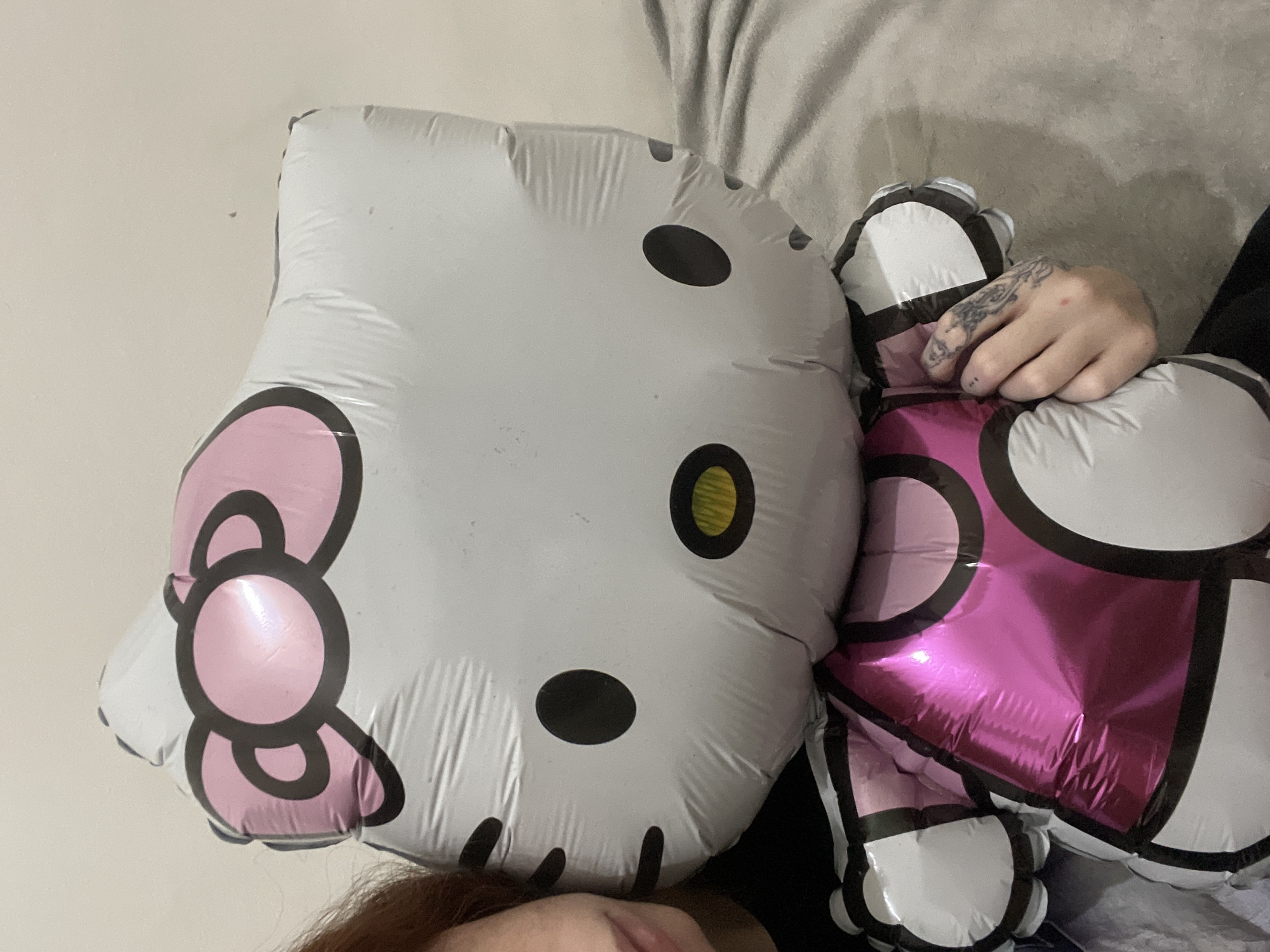 A close-up shot shows a person holding two partially deflated Hello Kitty foil balloons. One balloon shows Hello Kitty's face with a pink bow, while the other shows Hello Kitty's body with a pink dress. A tattooed hand rests on the body balloon. The background includes a white wall and a grey, possibly wrinkled, fabric. Part of the person's red hair and mouth are visible at the bottom of the frame. The location of the scene cannot be determined from the given image.