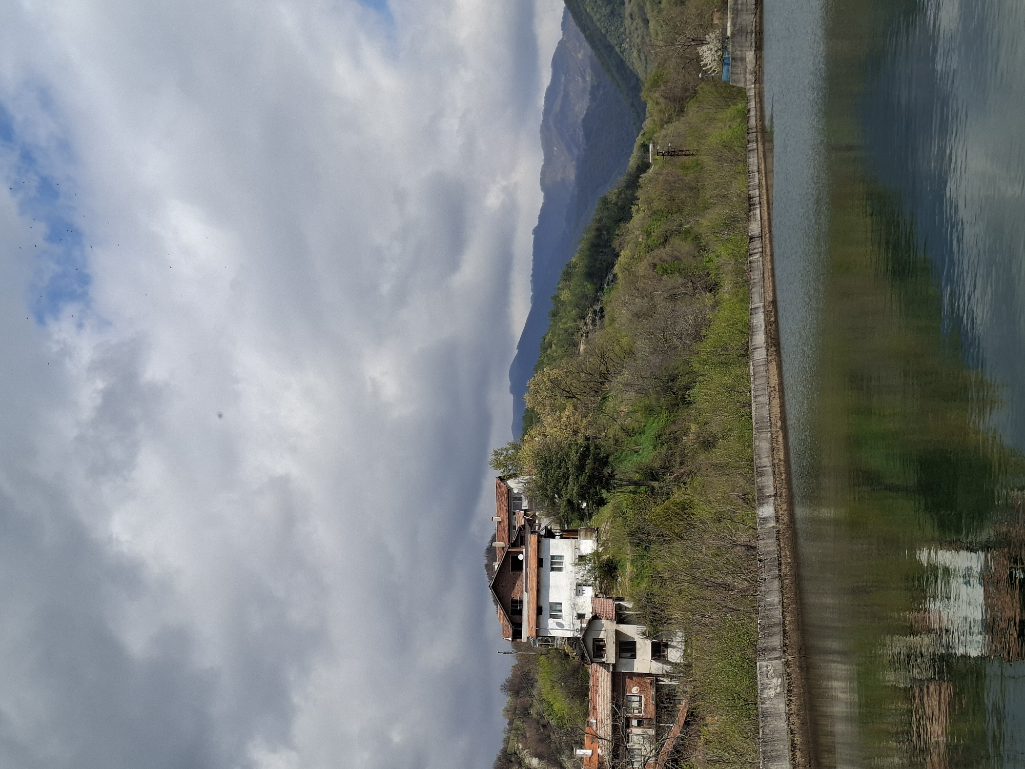 The image depicts a landscape featuring a body of water, possibly a lake or river, with reflections of the surrounding environment. A concrete wall runs along the edge of the water. Adjacent to the wall is a hillside with vegetation and several buildings, including houses with red-tiled roofs and white walls. In the background, there are mountains partially obscured by clouds. The sky is overcast with a mix of white and gray clouds. A flock of birds is visible in the sky. The image appears to be taken from a slightly elevated perspective.