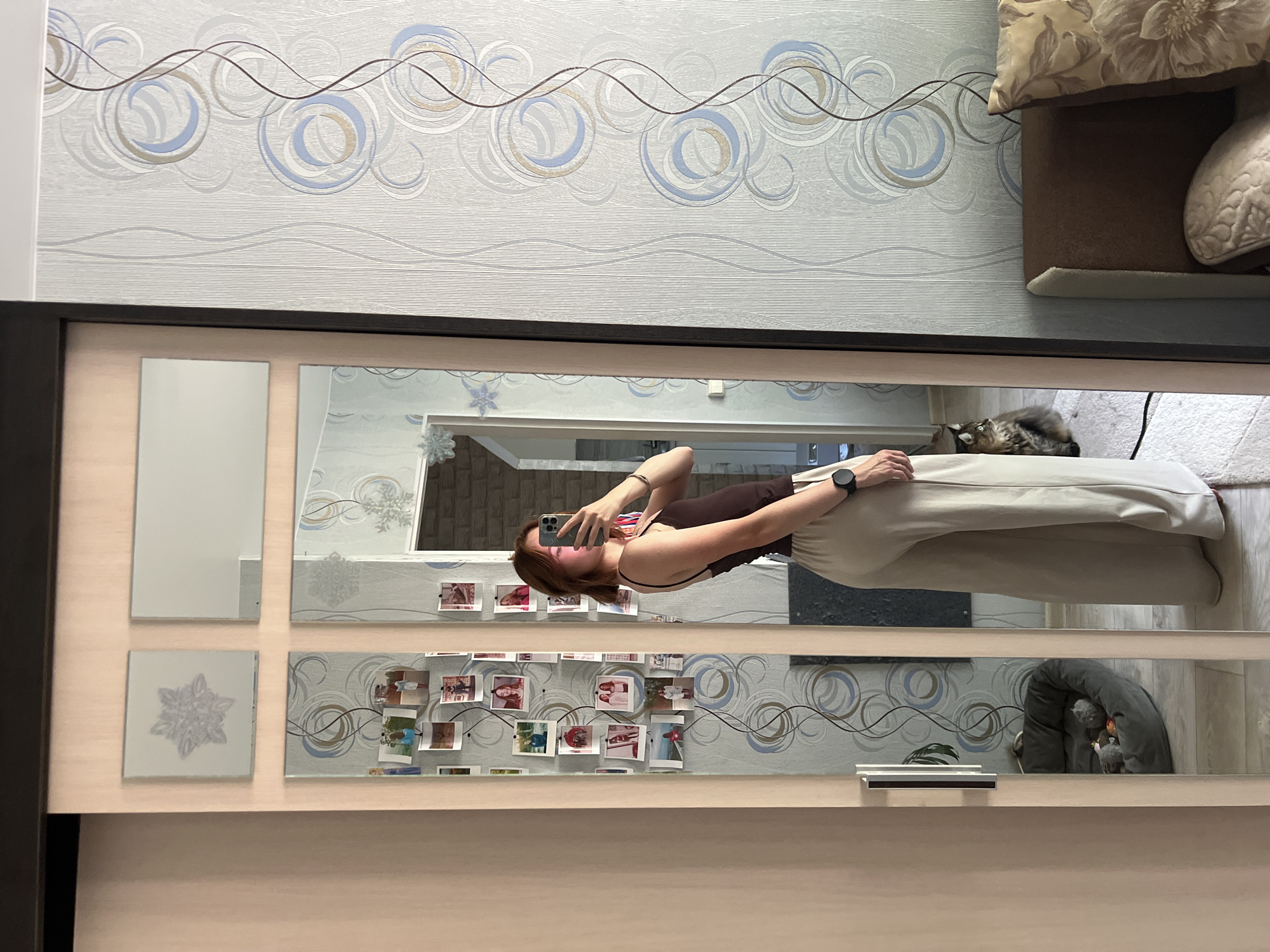 A young woman with shoulder-length brown hair is standing in front of a mirrored wardrobe, taking a selfie with her smartphone. She is wearing a dark brown tank top and loose, beige pants. She is looking at her reflection in the mirror. To her left, the wardrobe door is partially open, revealing a light-colored closet interior. The mirrored surface of the wardrobe is divided into four panels, decorated with various printed images, including several photographs of people and some snowflake designs. On the right side of the image, a patterned wallpaper with swirling blue and tan circles is visible. A cat is sitting on the floor to the woman's right, facing away from the camera and looking to the side. A dog bed with toys is reflected in the lower left mirror panel. A part of a bed with decorative pillows is visible on the right side of the image. The scene appears to be an indoor residential setting.