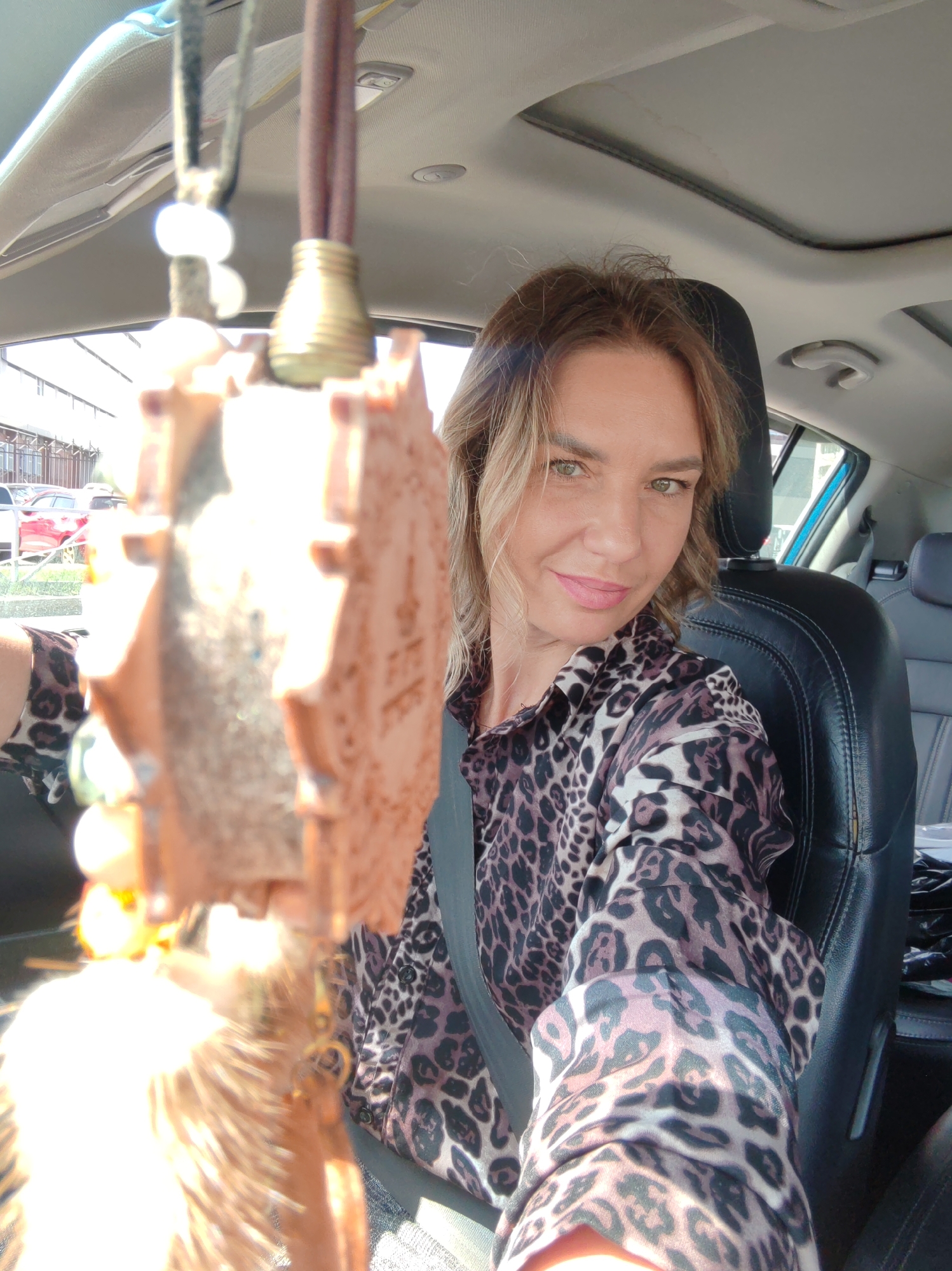 A woman with light brown hair and green eyes is captured in a selfie-style shot from inside a car. She is wearing a brown and black leopard print long-sleeved shirt and has a seatbelt fastened across her chest. A gentle smile plays on her lips, accented by light pink lipstick.
In the foreground, hanging from the rearview mirror, is a large, out-of-focus ornament, possibly carved from wood, with a rustic, blocky design that might depict an animal or tribal symbol. It features beads and what appear to be feathers at its lower end, brightly illuminated by sunlight.
The interior of the car features black leather seats and a grey headliner with a visible sunroof. Through the car windows, an outdoor scene is discernible, showing buildings, fences, and parts of other vehicles, including a red car, indicating an urban or commercial setting. The strong sunlight suggests it is a bright, sunny day. The woman appears to be in a pleasant mood. No discernible text is visible in the image. The location is Petropavlovsk-Kamchatsky, Russia.