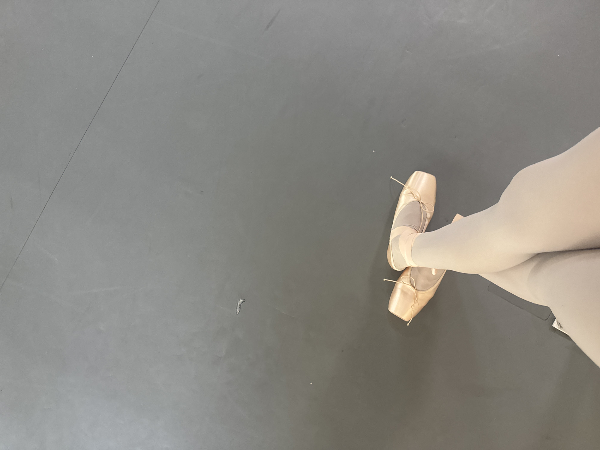 The image shows a dancer's legs and feet in ballet pointe shoes and tights, standing on a gray floor. The dancer's legs are crossed at the ankles. The pointe shoes are light pink. The floor appears to be a studio floor.