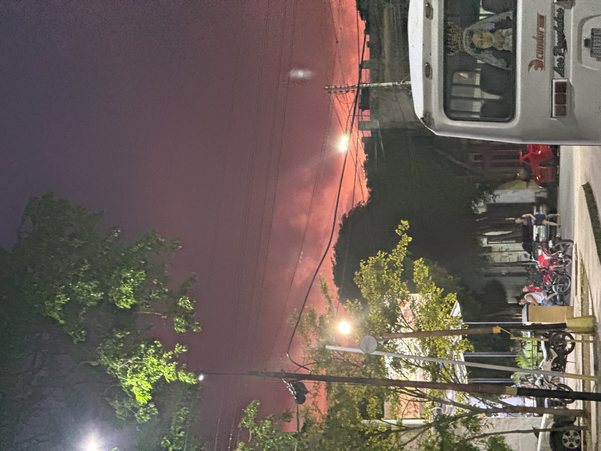 The image depicts a street scene at dusk or night, characterized by a prominent reddish-orange sky, suggesting either a sunset or atmospheric conditions like smoke. In the foreground, a white bus is parked on the right side, its rear visible. Through the rear window of the bus, a statue of a crowned figure, possibly a religious icon, is visible. The bus has text on its back, including "condor" and "Los Pugala".
To the left of the bus, a street extends with parked motorcycles and a car. Several people are present, some sitting near the motorcycles, others walking in the background. Trees with green foliage line the street, and streetlights are visible, casting a warm glow. Electrical wires are strung across the sky, and utility poles are also present.
In the background, buildings with architectural features resembling battlements can be seen. The overall environment appears to be an urban or semi-urban setting. The specific location in Santa Ana, Venezuela, is not explicitly indicated by any visual cues in the image.