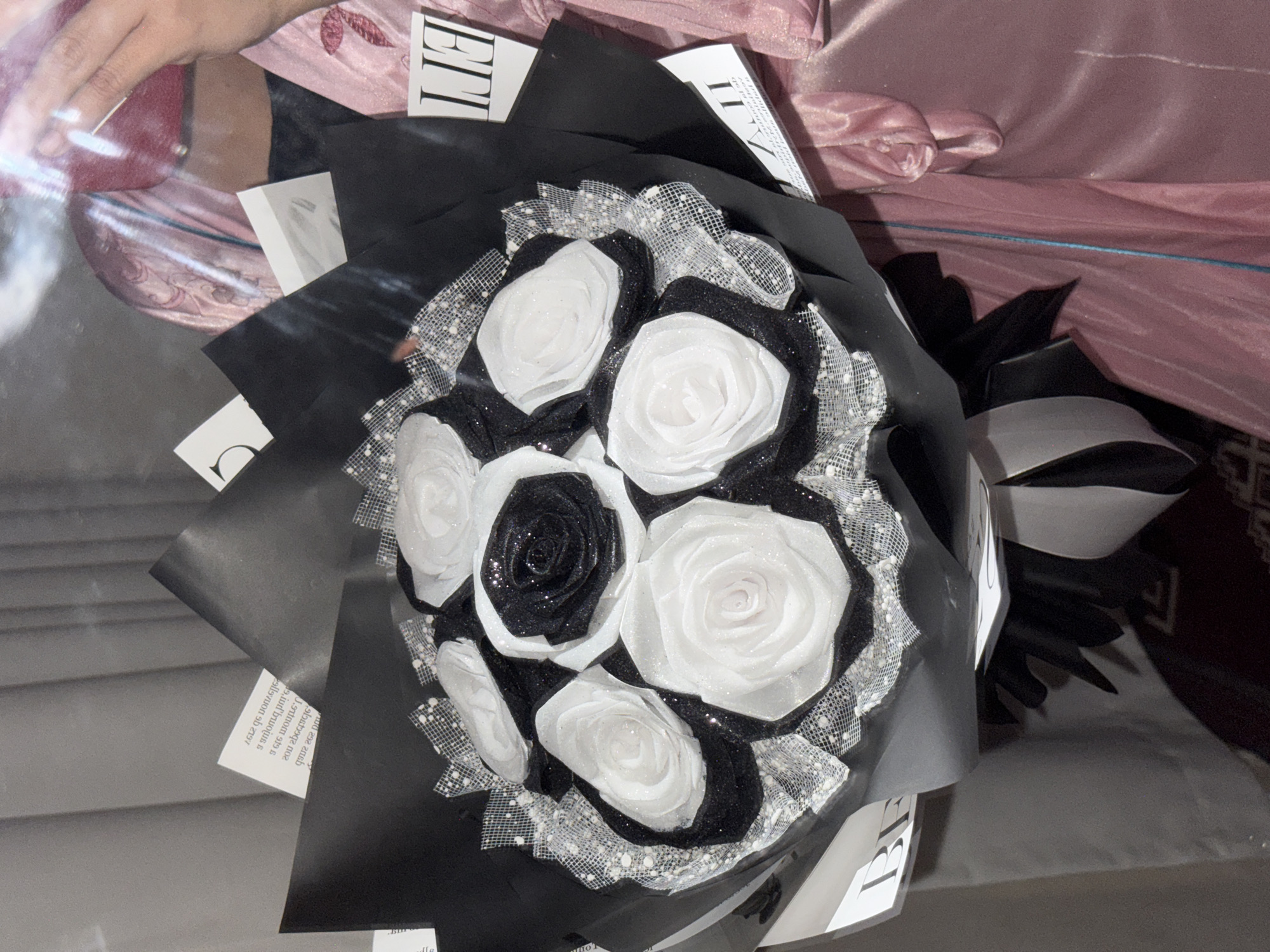 The image depicts a bouquet of artificial flowers, predominantly white with one black rose, wrapped in black paper and accented with glittery netting. It is positioned indoors on a surface that appears to be a bed or similar cushioned area, given the pink, silky fabric in the background. There is a hand in the upper left corner of the image. The surrounding environment includes printed materials with text, possibly from magazines or newspapers, which are incorporated into the bouquet's presentation. The scene suggests a staged or arranged setting rather than a naturally occurring one. The location in Davao City, Philippines is not discernible from the visual information in the image.