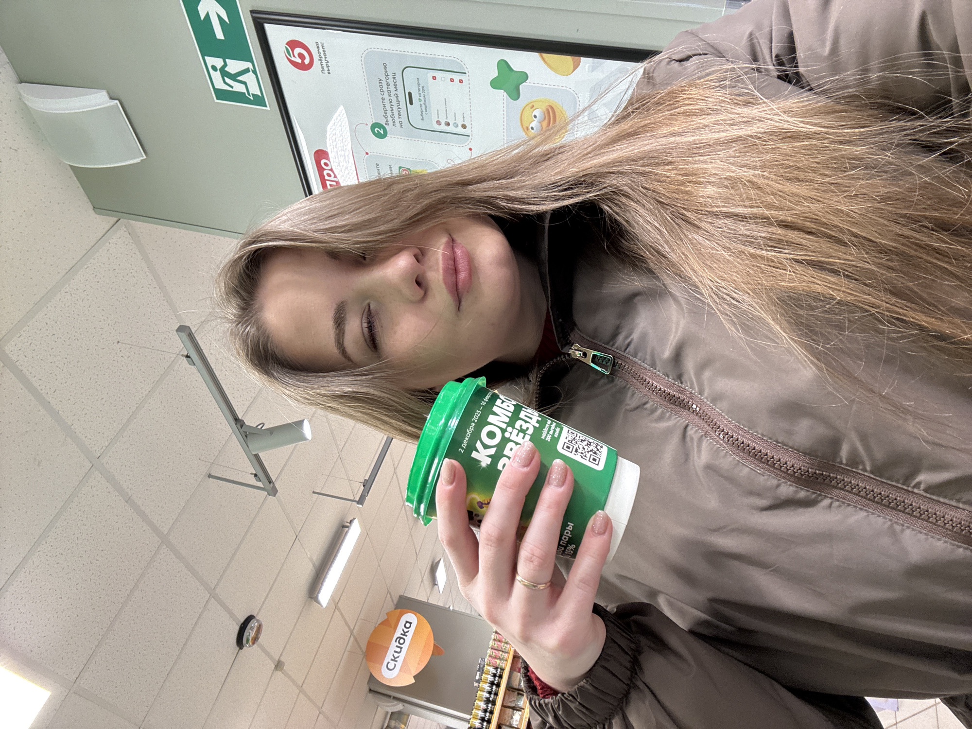A young woman is depicted in a selfie taken inside a Pyaterochka supermarket in Chaikovsky, Russia. She has long, light brown hair and is wearing a dark brown zippered puffer jacket over a reddish garment. She holds a green and white disposable coffee cup in her left hand, which bears the Russian text "КОМБО ЗВЕЗДА" (Combo Star), a QR code, and dates "2 декабря 2023 - 14 февраля 2025". A simple gold band ring is visible on her left ring finger. Her left eye is partially closed, and her lips are slightly pursed. The background features typical supermarket elements: a white acoustic tile ceiling with fluorescent and track lighting fixtures, and a circular smoke detector. To the left, shelves stocked with products are visible beneath an orange circular sign displaying "СКИДКА" (DISCOUNT). To the right, a large promotional poster for "Пятёрочка" (Pyaterochka), featuring a red '5' logo, text about selecting a loyalty category, and various emojis, is mounted on a light green wall. Above this poster, a green emergency exit sign with a white running figure and an arrow pointing right is present. The environment is uniformly lit by artificial lighting.