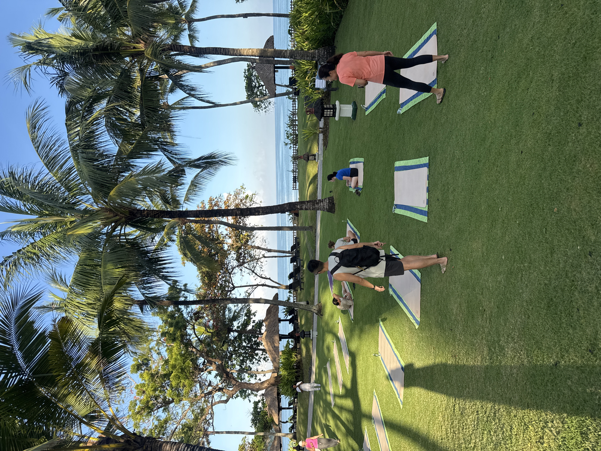 People are practicing yoga on a lawn in Jimbaran, Indonesia. The location is near the beach, with palm trees and other tropical vegetation. Some people are using mats. There are decorative lamps and a thatched-roof structure in the background. The sky is clear.
