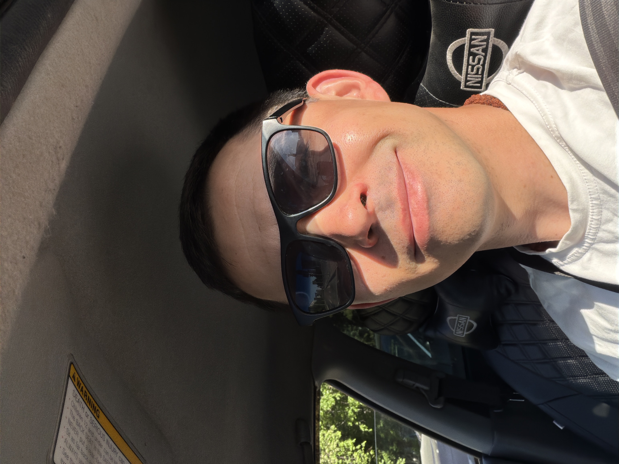 A man is photographed from a slightly low angle inside a car. He wears sunglasses and a white T-shirt. The interior features a Nissan seat cover. An "A WARNING" sticker is visible on the left. Outside the car window, there are trees. Location information (city of Izhevsk, Russia) is not discernible from the image.