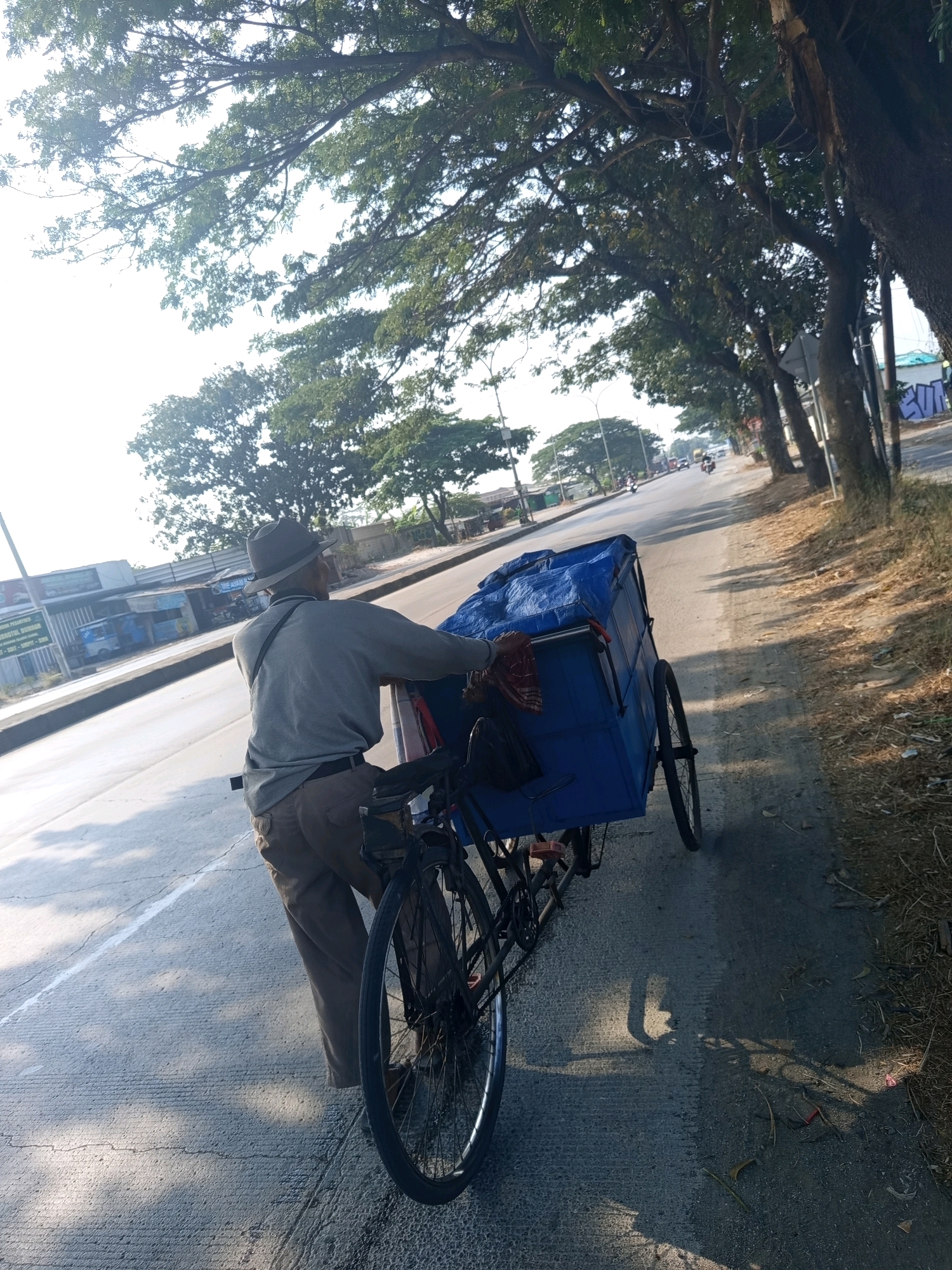 A man wearing a hat and a grey long-sleeved shirt is pushing a bicycle-drawn cart with a blue tarp on the side of a road. The cart has two wheels in the back and the bicycle is attached to the front. The man is walking on the shoulder of the road, which is lined with trees. In the background, there are buildings and other vehicles on the road. The sun is shining, casting shadows on the road. The scene appears to be a street in Karawang, Indonesia.
