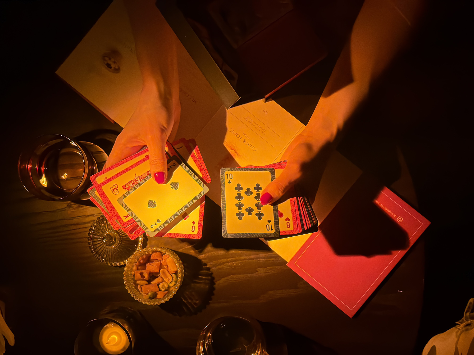 The image presents an intimate, dimly lit scene, likely set in a bar or lounge in Đà Nẵng, Vietnam, during the evening. The warm, artificial light, possibly from candles, casts a golden glow and strong shadows across a dark wooden table.
At the center, two sets of hands, with bright pink nail polish, are engaged in playing cards. One hand is holding a fanned-out hand, revealing cards such as a "King of Spades" and a "2 of Spades" from a deck with an ornate red and gold backing. The other hand is in the process of dealing cards, with a "10 of Clubs" visible on the top card of the deck being held.
Various objects related to a relaxed social setting are scattered around the table. To the left, a glass holds an amber-colored drink, reflecting the soft light. Nearby, a small, ornate glass bowl contains a mix of snacks, including nuts and dried fruit. A lit candle in a metallic holder further contributes to the warm ambiance.
In the background, an open, pale-colored box or menu displays text including "CHAI & TONIC INDIA," "GIN MASALA CLAR TEA," and "IDERFLOWER-TONIC," along with the number "220." A red, rectangular item, possibly an envelope or a coaster, bears the text "thekeepsakebar.com" and an Instagram logo, suggesting the establishment's name. The overall scene evokes a sense of cozy leisure and entertainment.
