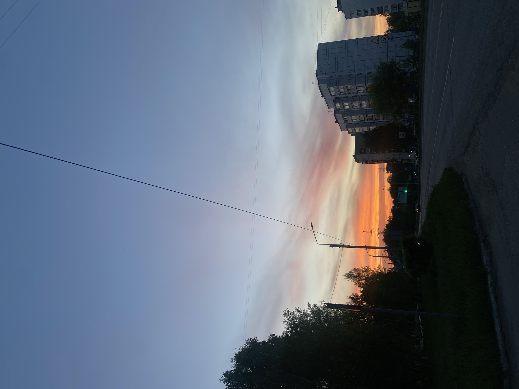 The image depicts a street scene in what appears to be a residential area of Chaikovsky, Russia, during a vibrant sunset. On the right side of the frame, there is an asphalt road with a curb. Adjacent to the road is a line of trees and dense foliage, partially silhouetted against the sunset. A street lamp is visible among the trees. Beyond the trees, there are several high-rise apartment buildings with multiple windows. The sky is a gradient of colors, ranging from a deep blue at the top to shades of orange, pink, and yellow near the horizon. Two parallel electrical or telephone wires stretch across the frame. A traffic light is visible with a green light showing. There is a road sign with a speed limit sign posted in front of a multi-story building.