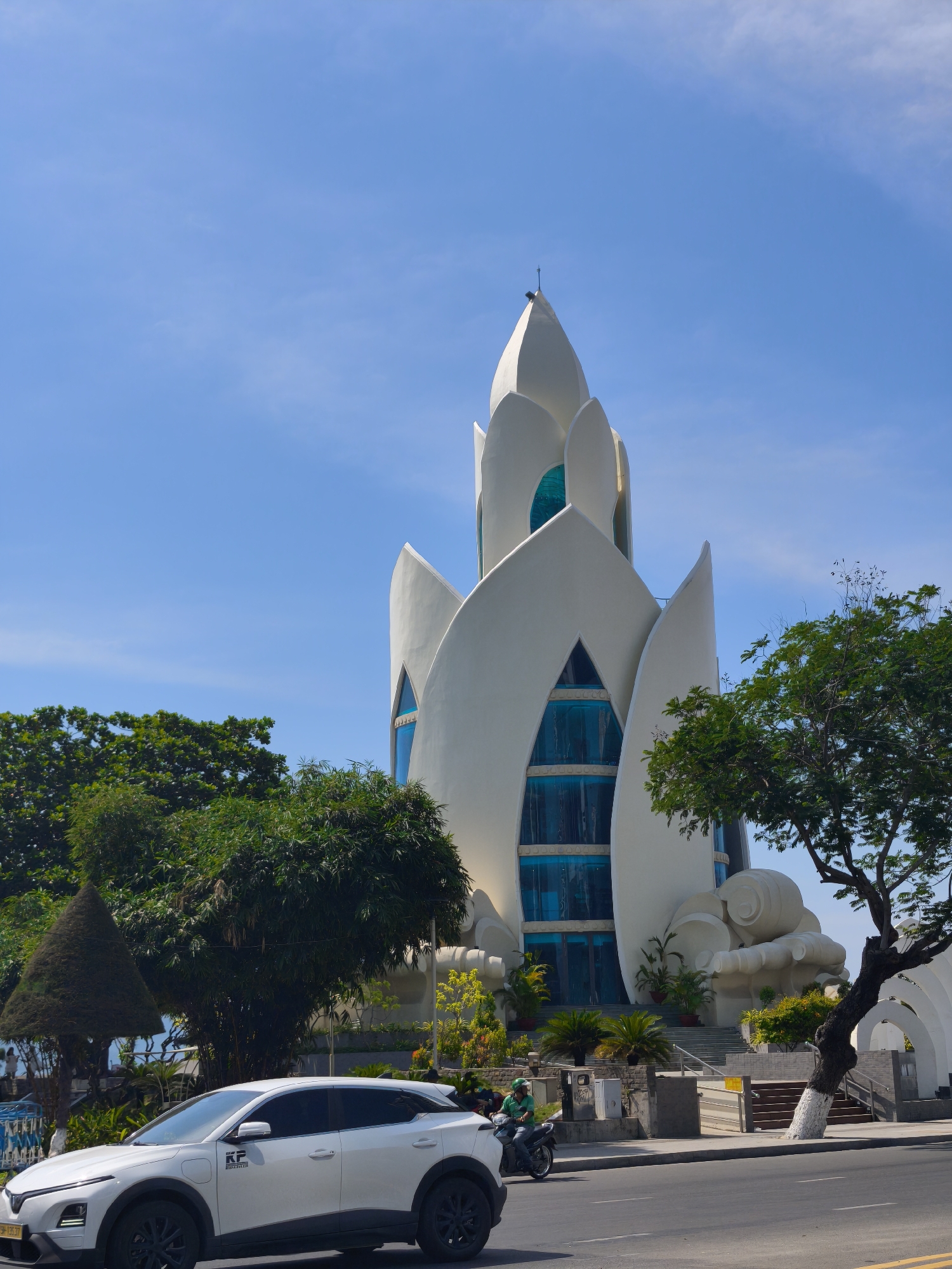 In Nha Trang, Vietnam, a white electric vehicle, possibly a VinFast VF e34, is parked on a paved road in front of a white, petal-shaped building with blue-tinted windows. The building's unique architecture resembles a blooming lotus flower and is situated amidst lush greenery. A person wearing a green shirt and a helmet is riding a motorcycle on the road to the left of the car. The sky is clear blue with a few wispy clouds. The overall scene suggests a bright, sunny day in a city known for its coastal attractions.