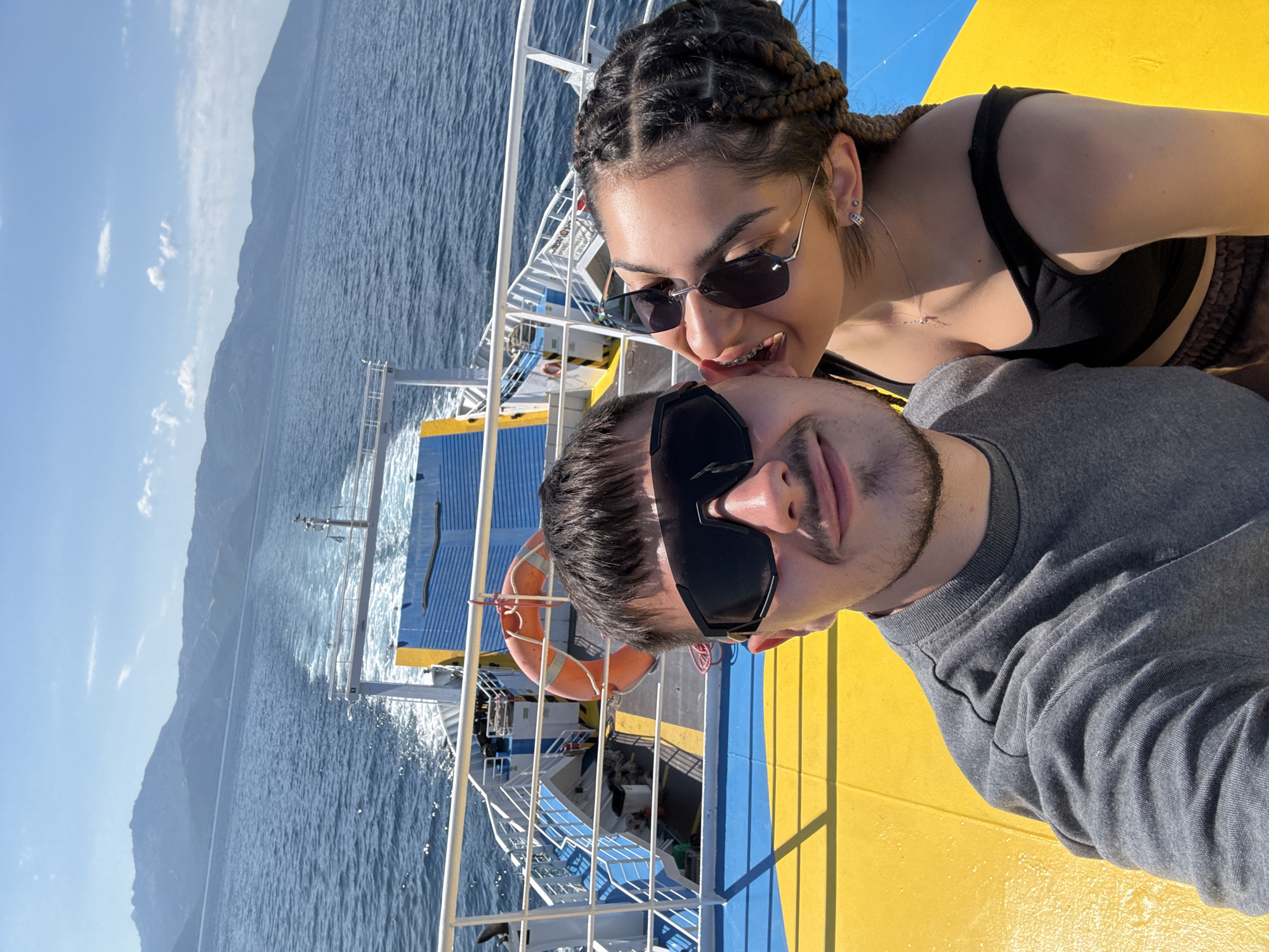 Two people are on the deck of a ferryboat in Greece, with mountains in the background. The person in the foreground on the left is a man wearing sunglasses and a grey t-shirt, taking a selfie. The person on the right is a woman with braided hair, also wearing sunglasses, leaning in to kiss the man's cheek. The ferryboat has yellow and blue painted decks and a railing. A life preserver is visible on the side of the boat. The water is blue, and the sky is clear with a few clouds.