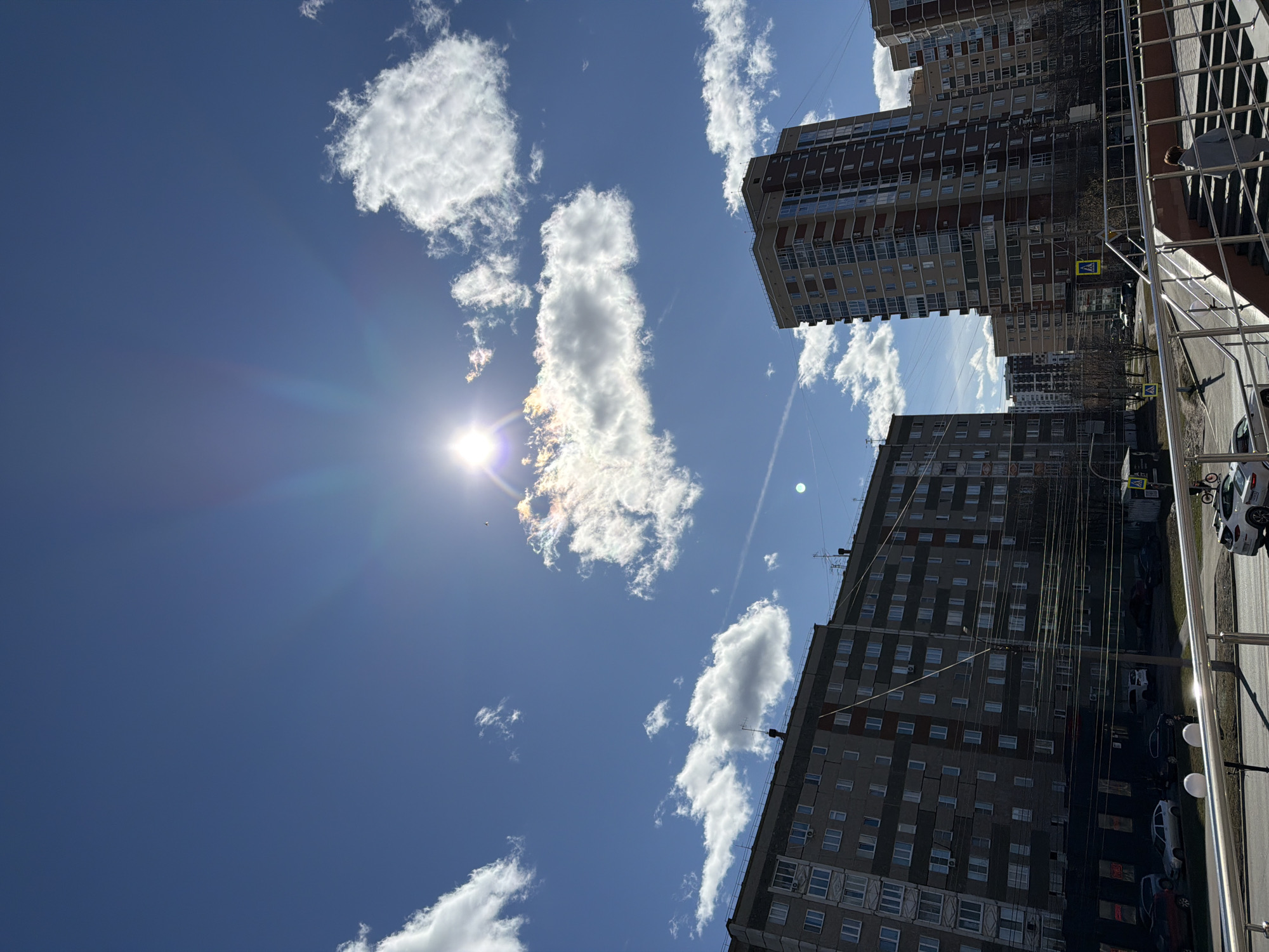The image captures a view from what appears to be a balcony or elevated position in a densely built urban area, likely in Izhevsk, Russia. The sky dominates the scene, featuring a clear blue backdrop interspersed with puffy white cumulus clouds. The sun is shining brightly, causing a glare and starburst effect. Below the sky, are tall apartment buildings with multiple floors and windows. A portion of a metal railing and a person are visible in the right portion of the image. Vehicles are parked alongside the base of the buildings.