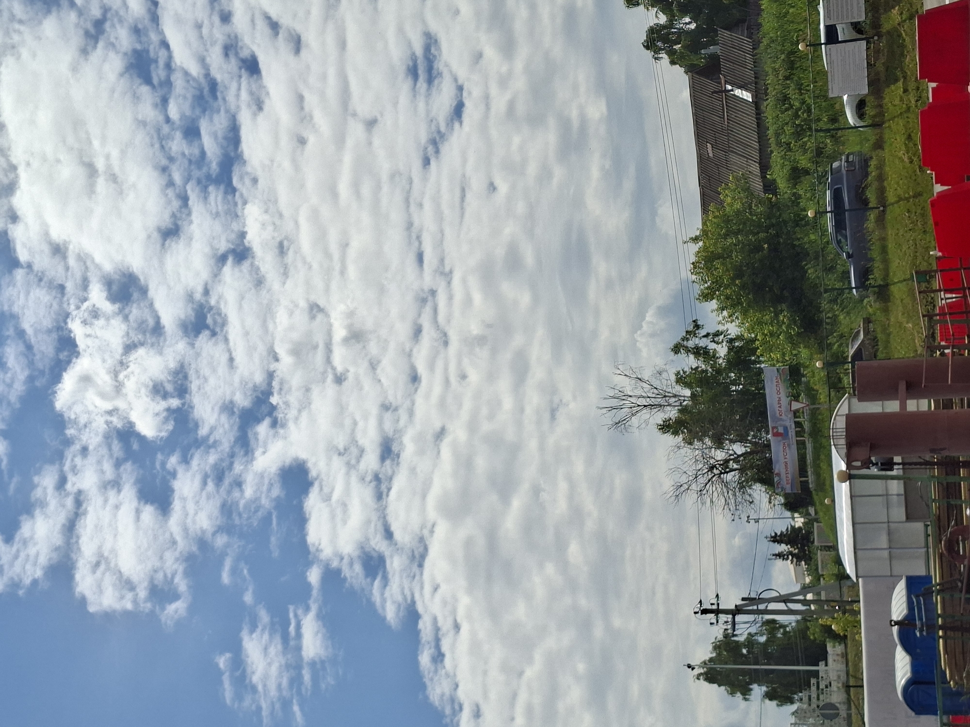 This is a vertically oriented, outdoor photograph taken during the daytime. The scene depicts a predominantly cloudy sky with patches of blue visible. The clouds are white and appear to be thin and wispy, suggesting a partly cloudy or overcast day.
In the background, there is a collection of buildings and trees. One building has a corrugated, weathered roof and appears to be older. Adjacent to it is a more modern structure with a white, reflective roof. A dark-colored car is parked on a grassy area. There are also several utility poles with wires crisscrossing overhead, indicating the presence of infrastructure.
To the right of the frame, there are rows of red stadium-style seats. Further into the scene, there are industrial-looking structures, possibly tanks or pipes, with a reddish-brown hue. A blue portable toilet is also visible in the foreground.
A banner with text is displayed on a white background, stretched between two poles. The text appears to be in Cyrillic script. One section of the banner shows what looks like a logo with red, white, and blue elements. The text is not entirely legible due to the image resolution and orientation. However, based on the provided location context, "Verhnij Uslon, Russia," it's plausible the banner contains information relevant to this area.
The overall impression is of a rural or semi-industrial setting, possibly near a public area or a construction site, under a cloudy sky. There are no people visible in the photograph. The scene does not convey any specific emotion and appears to be a straightforward documentation of the environment.
