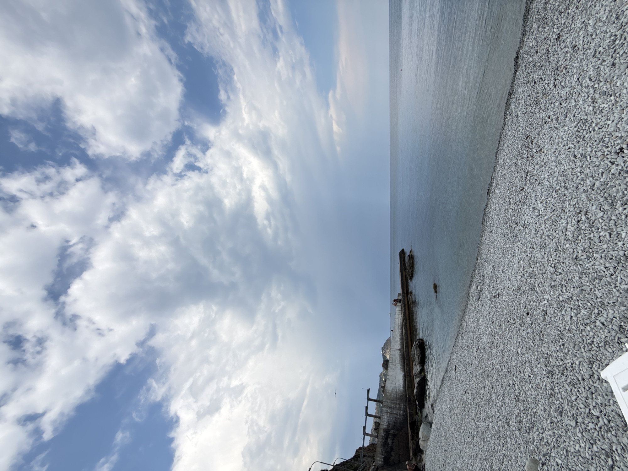 The image depicts a pebble beach with a calm sea under a partly cloudy sky. In the background, a concrete pier extends into the water, with a few people visible on it. A weathered concrete structure with supporting pillars is present on the left side of the beach, adjacent to a rocky cliff. The sea is a muted blue-grey, and the sky features a mix of bright white clouds and darker, more overcast areas, with shafts of light breaking through. A single bird is in flight near the concrete structure. The overall scene suggests a coastal location, potentially a less developed or more natural beach area. The image was taken in the city of Partenit, Ukraine.
