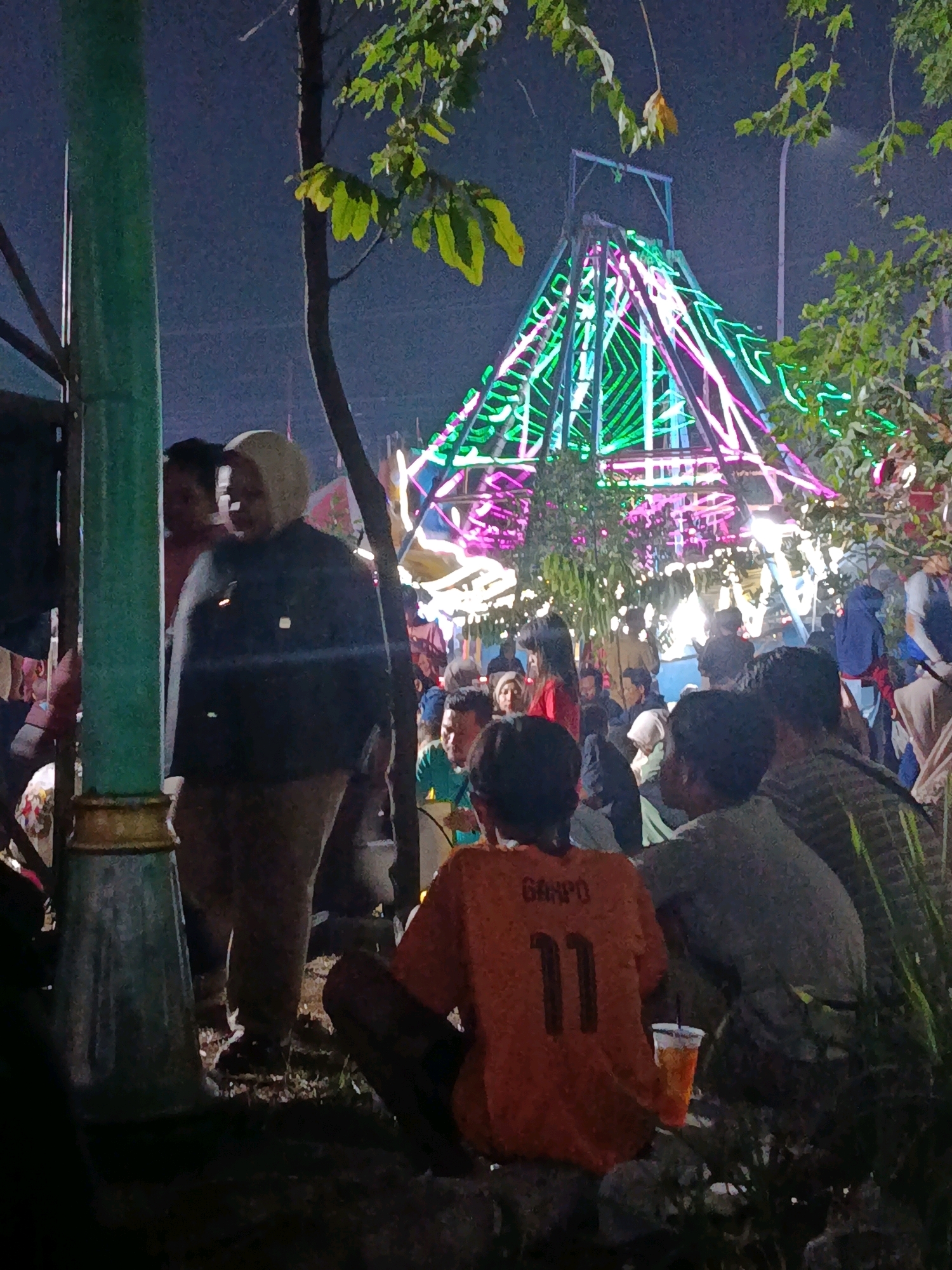The image depicts a nighttime scene at a fair or carnival in Balaraja, Indonesia. In the foreground, several individuals are gathered, with a person in an orange t-shirt with the number "11" and "GARPO" visible on the back, sitting down. To the left, a green pole and tree trunk are prominent. In the background, a large, illuminated structure, possibly a Ferris wheel or similar ride, is decorated with green and pink neon lights. The environment appears to be outdoors, with a dark sky suggesting it is night. There are other people visible in the mid-ground and background, indicating a public gathering.