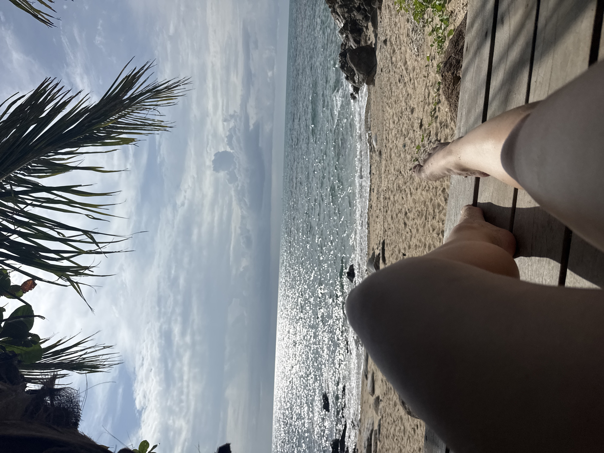 This is a first-person perspective image taken from a wooden platform at a beach location, likely in the Ko Pha-ngan District, Thailand. The foreground features the legs and feet of a person lounging on the platform, with the sand and ocean stretching out before them. Palm fronds hang from above, framing the top of the image and providing shade. The ocean is a deep blue, with sunlight glinting off its surface. The beach itself is sandy, with scattered rocks and some vegetation visible. The sky is partly cloudy, with dramatic formations of white and gray clouds against a blue backdrop. The scene suggests a relaxed and tropical atmosphere.