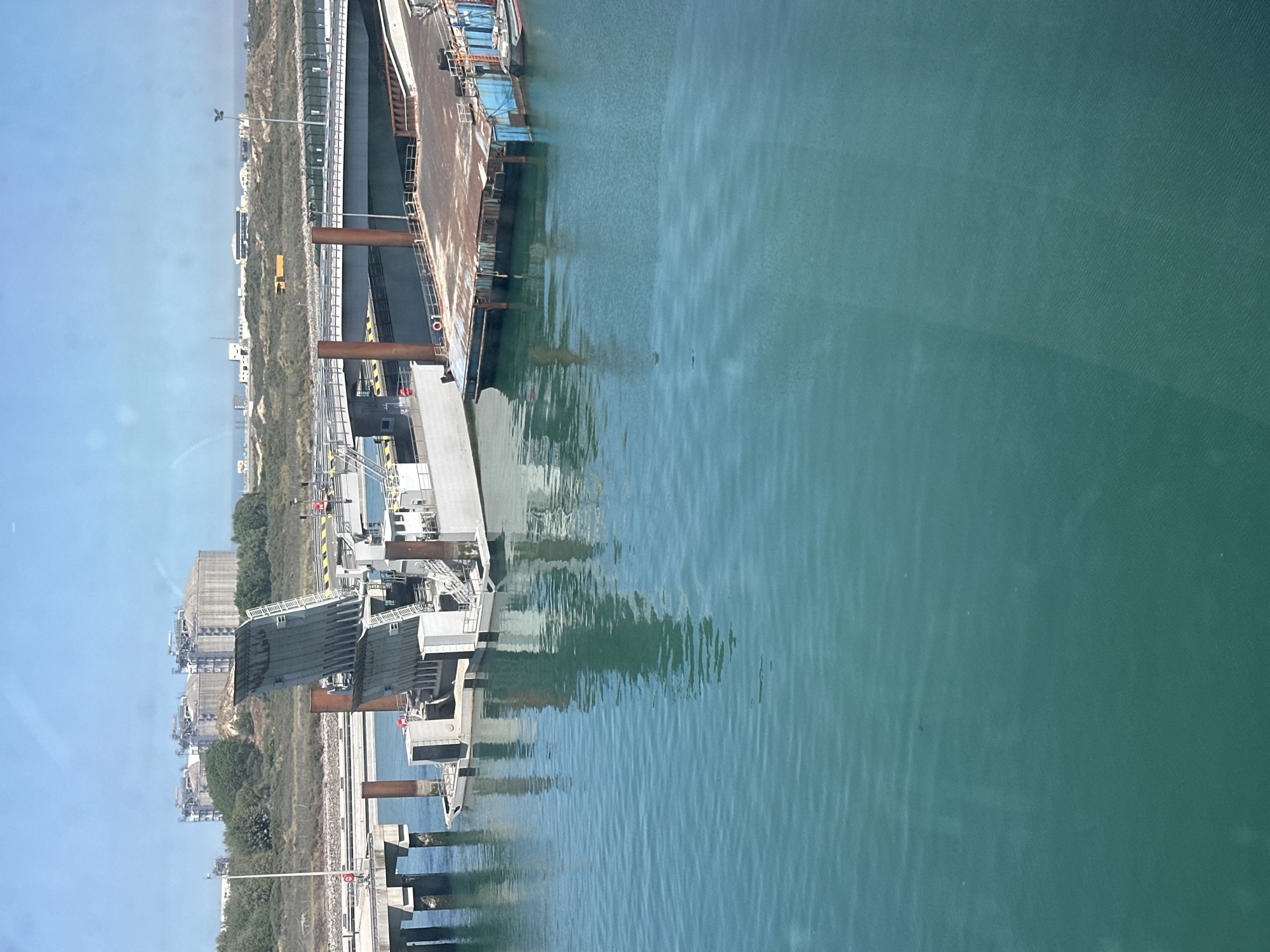 This image shows a dock and body of water in Loon-Plage, France. The photo captures the edge of a body of water, reflecting the structures and sky above. The structures appear to be industrial buildings and docks with metal beams and machinery. The water is a light green color, with ripples and reflections visible on the surface. The environment consists of an industrial area next to a body of water under a bright sky.