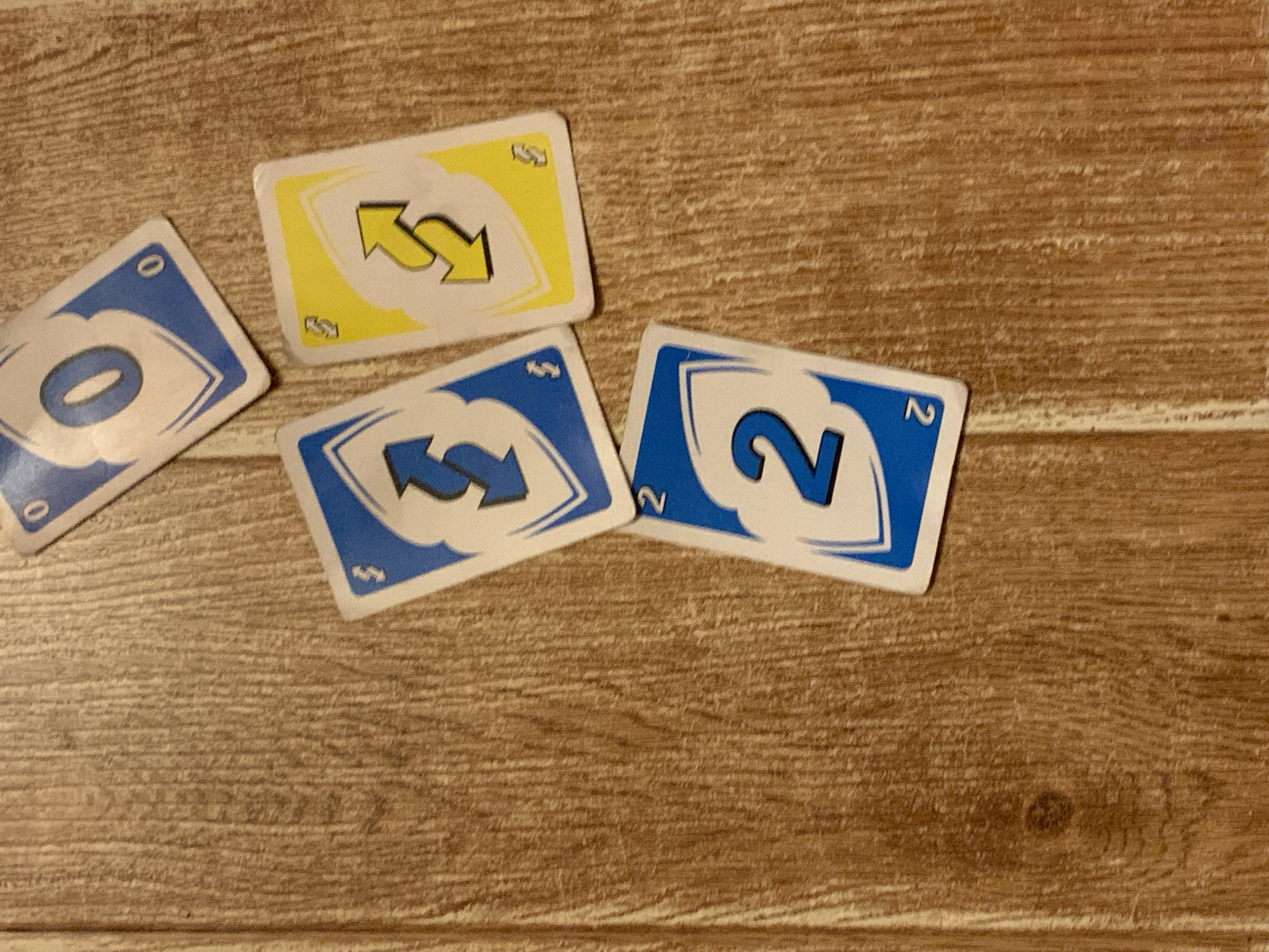 Four Uno cards are scattered on a wooden surface. The cards are as follows: a blue zero, a blue reverse card, a yellow reverse card, and a blue two. The wooden surface appears to be a table or floor. The lighting in the image is dim, suggesting it might be indoors. The location is not specified, but the context implies it's a casual setting for playing cards.
