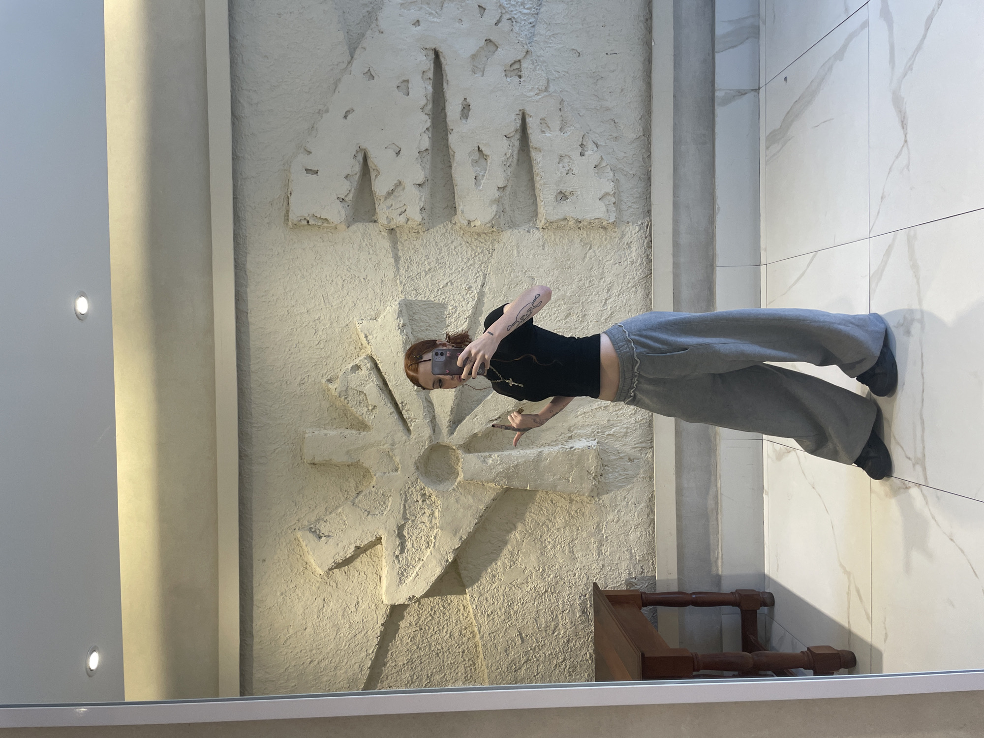 A mirrored image depicts a woman standing in what appears to be an interior space, likely a building or museum. She is positioned against a wall, taking a selfie with her mobile phone.
The woman has red hair and is wearing a black top and gray sweatpants. Her left arm is tattooed.
Behind her, the wall features decorative elements: large, textured, white letters "MAMA," and below them, a radiating circular design resembling a flower or a stylized sun.
To the right of the wall and the woman, the floor is tiled with white marble tiles. A wooden furniture piece (possibly a chair) is partially visible in the lower part of the reflected image.
The scene appears to be captured through a mirror, with the edges of the mirror frame visible. The mirror reflects other elements of the room, including a plain white wall with embedded lights. The location is in Buenos Aires, Argentina, identified by the decorative wall elements.
