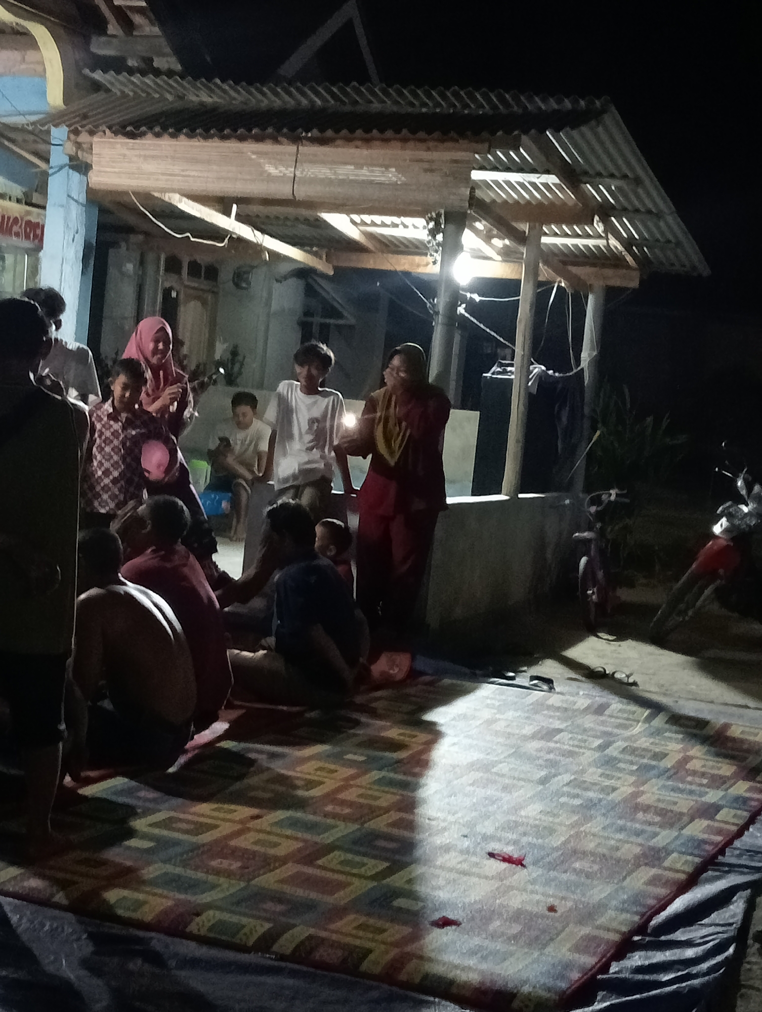 The image depicts an outdoor gathering at night, likely in Rasuan Baru, Indonesia. A group of people, including adults and children, are assembled around a covered outdoor area with a corrugated metal roof supported by wooden poles. A single light bulb hangs from the roof, providing illumination. A patterned mat is spread on the ground where several individuals are seated or kneeling, some of whom appear to be children. One person, wearing a pink headscarf, is holding a pink, translucent object. Another individual, dressed in a red outfit, stands near a concrete ledge. In the background, two motorcycles are parked, one red and one darker colored. The scene suggests a casual social event taking place outdoors after dark.