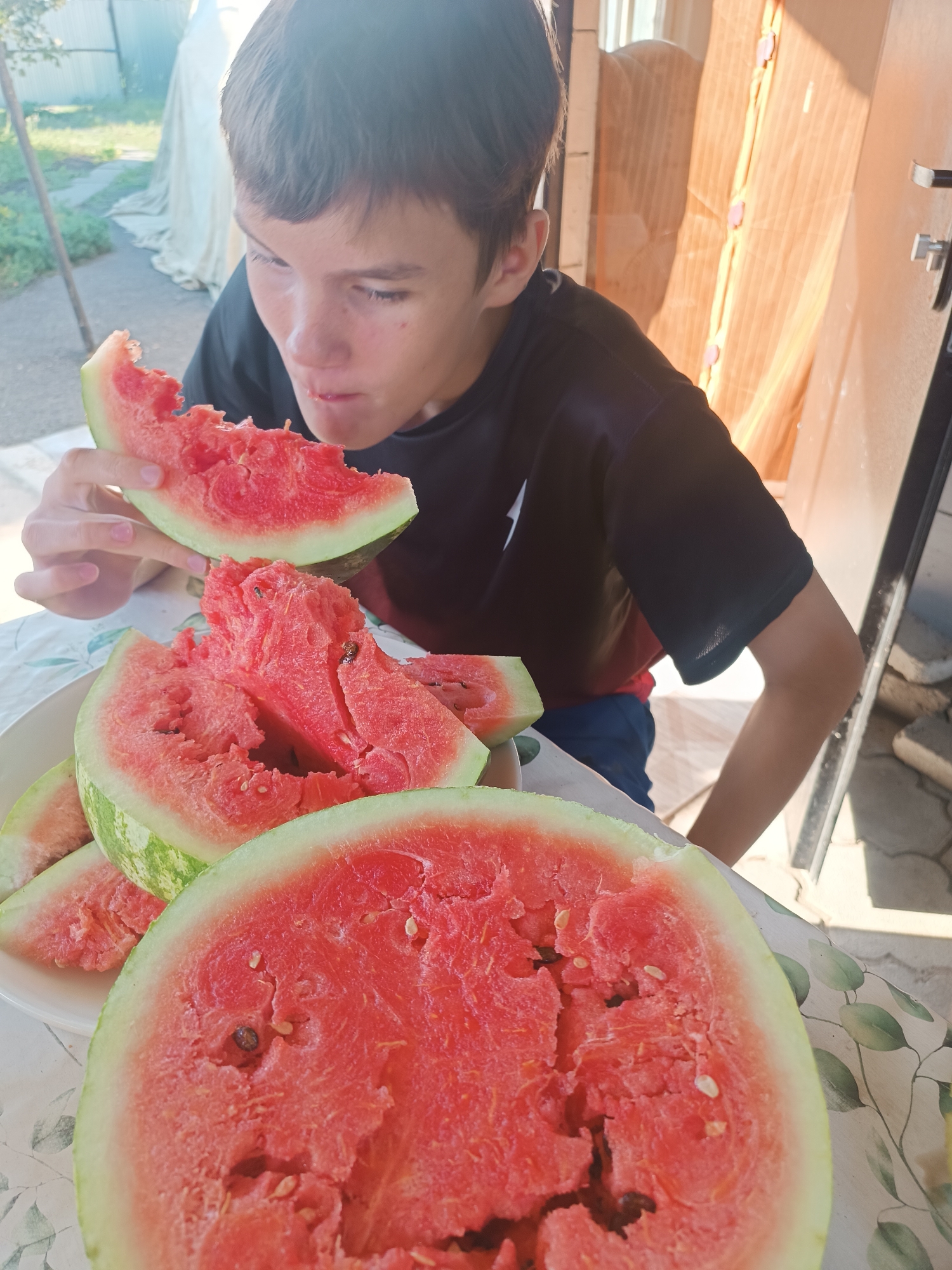 A young boy with brown hair, wearing a black t-shirt and blue shorts, is eating a slice of watermelon in an outdoor setting. He is holding a large slice of watermelon in his left hand and is bringing it to his mouth. Several other slices of watermelon are visible on a white plate in front of him, some of which have been partially eaten. The table is covered with a white tablecloth patterned with green leaves. In the background, there is a wooden structure and some green foliage, suggesting a residential area. The lighting indicates it is daytime. The location is Бөрлі ауылдық округі, Kazakhstan.