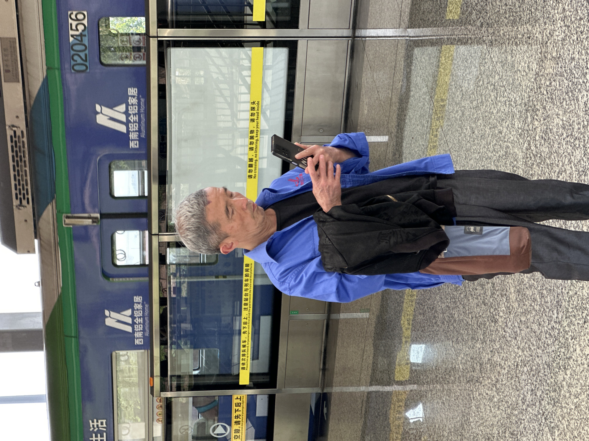 An elderly man with gray hair is standing at a subway station platform in Jiansheng, China. He's looking down and using a smartphone. He is wearing a blue button-down shirt over a black t-shirt, black pants, and a black jacket is slung over his shoulder. A bag with brown, grey and blue panels is also partially visible. Behind him is the subway car, marked with "020456" and the logo of "Southwest Aluminium Home". A sign on the subway door reads "No crossing, no littering, keep your head inside". The ground of the platform is a speckled grey.