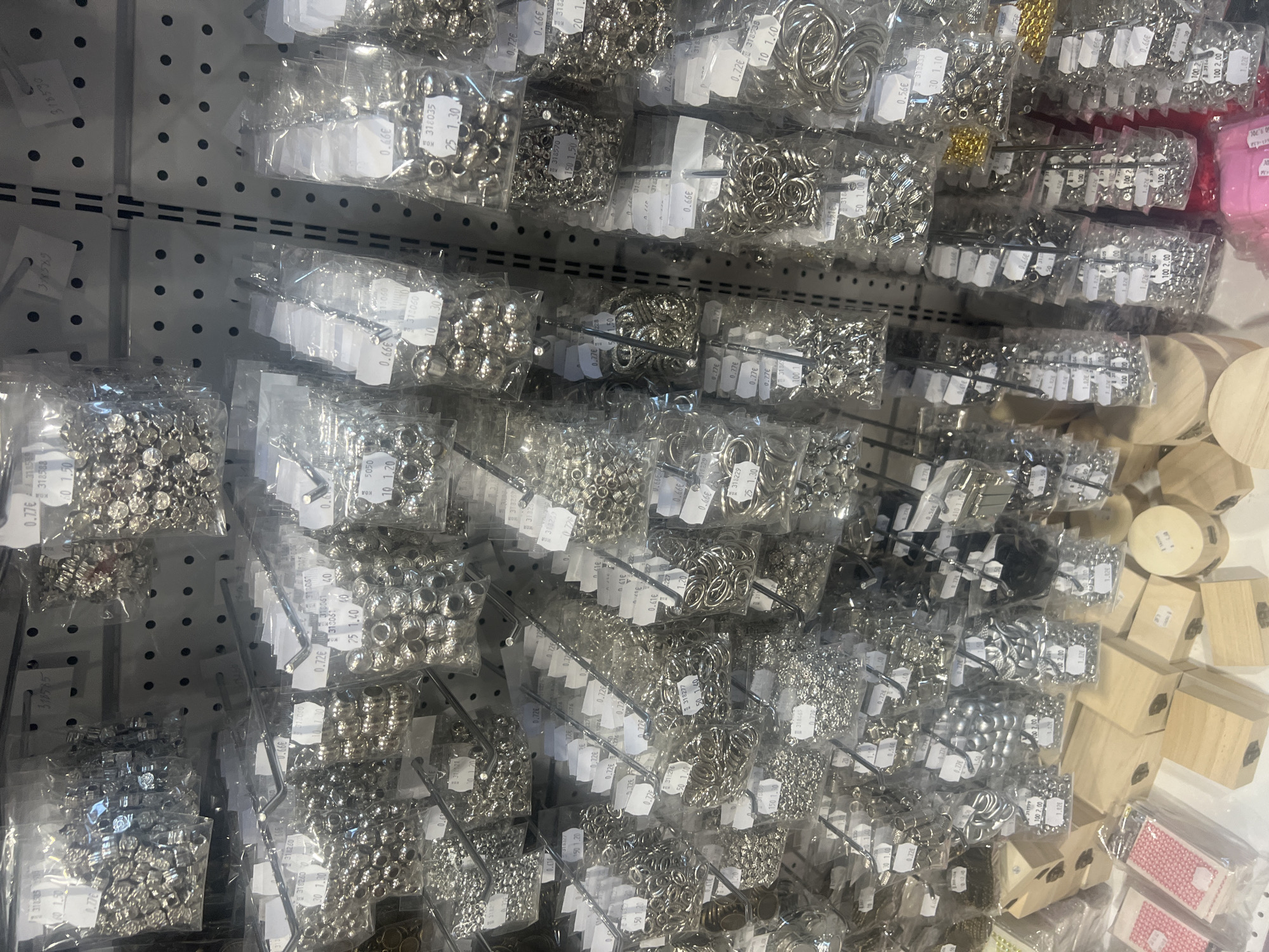 The image shows a close-up view of a wall display inside a store, likely a craft or hobby shop. The display consists of numerous small, clear plastic bags filled with various crafting components, such as beads, metal rings, and other embellishments. Each bag is individually hung on a metal grid, which is part of the store's shelving system. Small white labels with prices and item codes are attached to each bag. Some wooden boxes and other packaged items are also visible on the shelves towards the right side of the image. The image likely was taken in the city of Burgas, Bulgaria.