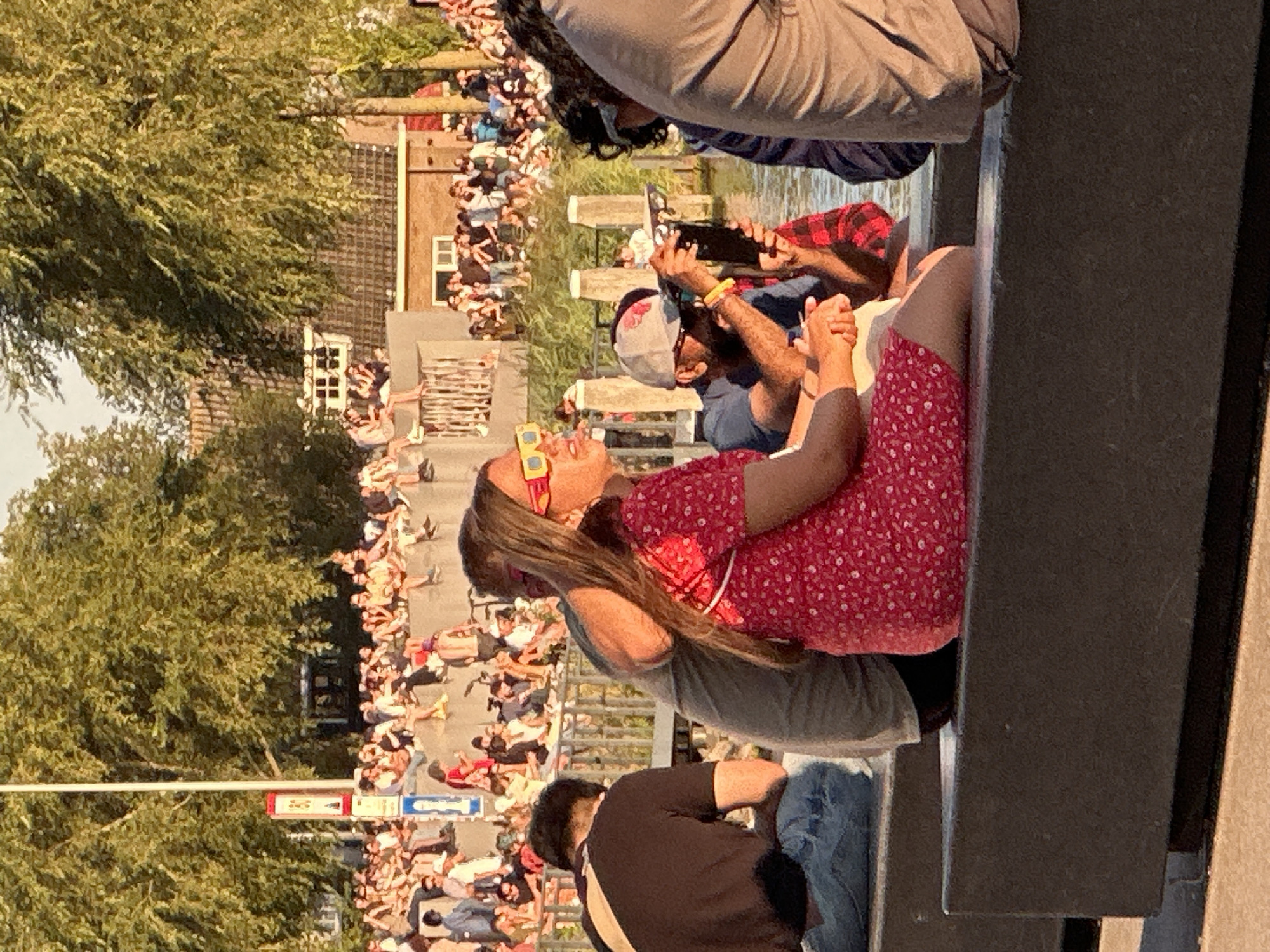 The image depicts a scene along a canal in Amsterdam, Netherlands. In the foreground, three individuals are seated on a dark-colored concrete structure. The person on the left is wearing a black t-shirt and blue jeans. Next to them, a person is wearing a grey t-shirt and appears to be leaning on the person in the red dress. This person is wearing a red floral dress and bright yellow-orange sunglasses. The person on the right, partially visible, is wearing a blue and white striped top. Behind these individuals, a man wearing a baseball cap and sunglasses is holding a dark-colored mobile device, possibly taking a photo or video. A crowd of people is visible in the background, gathered along the canal bank and on elevated structures, suggesting a public event or gathering. Trees with green foliage line the canal, and buildings with tiled roofs are visible in the distance. The lighting indicates it is daytime, likely late afternoon or early evening, with the sun casting long shadows. The presence of boats and water is implied by the canal setting.