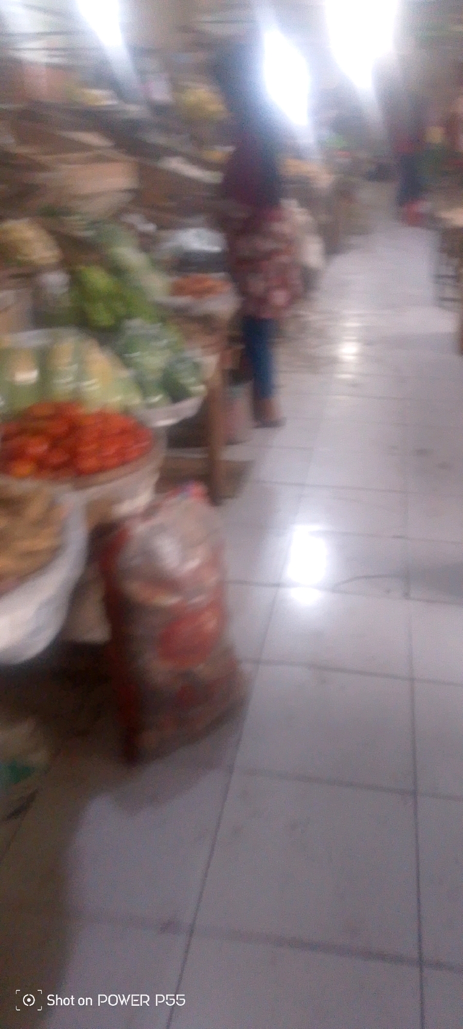 The image appears to be taken inside a market. In the foreground, there are some blurred items, possibly produce. To the left, there's a pile of red items, likely tomatoes, in a container. To the right, a person is visible, wearing a patterned shirt and blue pants. The floor is tiled. There are also some bags, one of which is translucent and appears to contain red items. The lighting is bright, suggesting indoor lighting or a well-lit area. The overall impression is of a bustling market scene.
