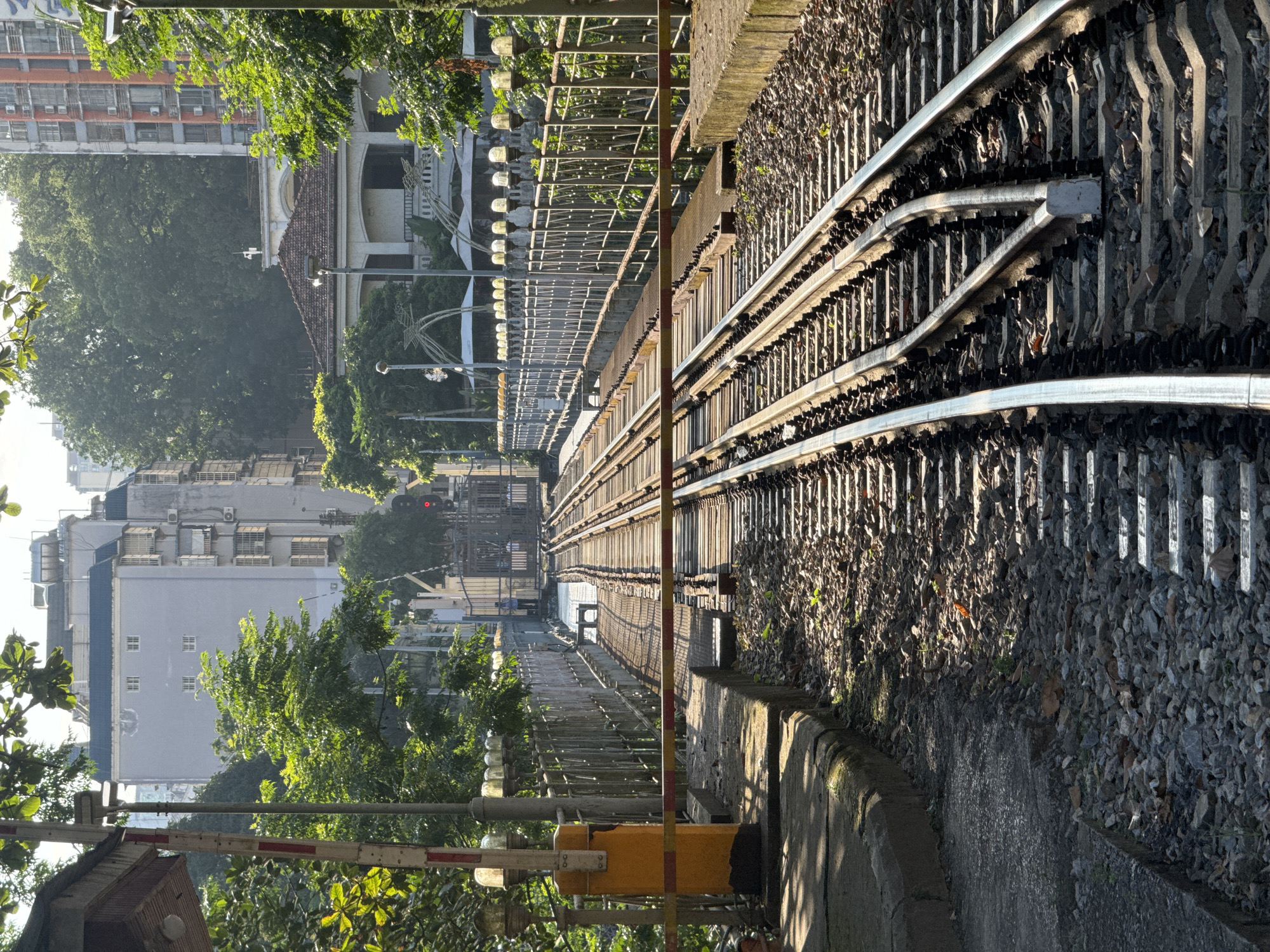 The image shows a railroad track in Lao Cai Ward, Vietnam. The tracks are surrounded by gravel. To the left, a pedestrian walkway is lined with a metal fence. Beyond the fence are buildings and trees. A yellow and red railroad crossing barrier is visible in the foreground on the left. The tracks converge in the middle distance.