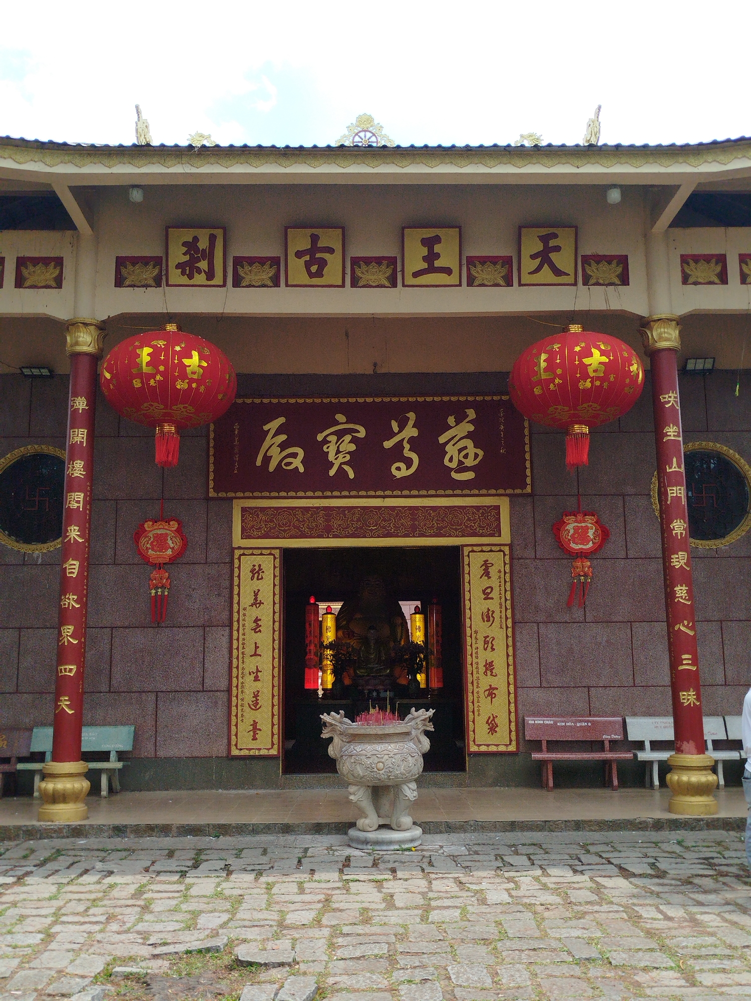 The image shows the entrance to a temple, possibly in Da Lat, Vietnam. The temple entrance has traditional architectural features, including a tiled roof, red pillars with gold accents, and prominent Chinese characters inscribed on panels above the entrance and on the pillars. Red lanterns hang on either side of the doorway, with decorative ornaments hanging below the lanterns. An incense burner sits in front of the entrance. Benches are placed on either side of the entrance. The ground in front of the temple is paved with stone. The image is taken during the daytime. The location of the temple is not apparent.