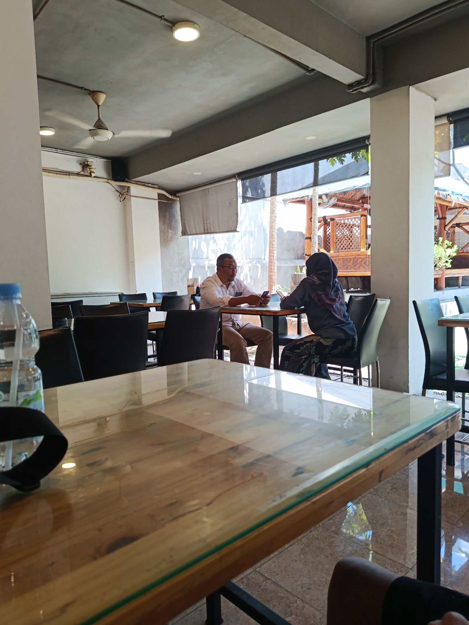 The image depicts the interior of a restaurant or cafe in Surabaya, Indonesia. In the foreground, a table with a wooden top and a reflective surface is prominent, with a portion of a black strap lying on it. To the left, a bottle of water is partially visible. In the middle ground, two individuals are seated at a table. A man, wearing a white shirt and light-colored trousers, is on the left, holding a mobile phone. A woman, dressed in a dark headscarf and patterned trousers, sits opposite him, also looking at her phone. The restaurant appears to have an industrial-style ceiling with exposed pipes and a fan. Other tables and chairs are visible in the background, along with windows that offer a glimpse of an outdoor area with bamboo structures. The flooring is tiled. The lighting suggests it is daytime.