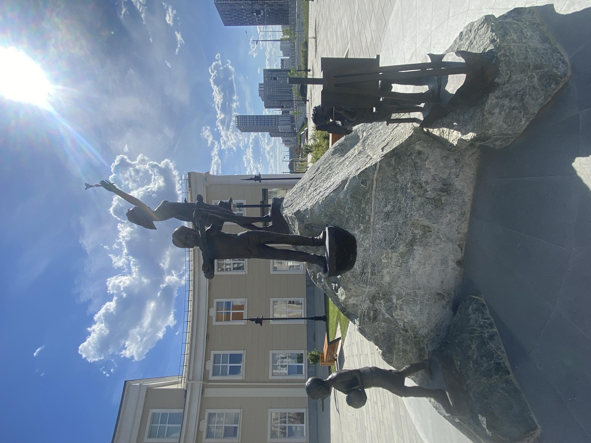 The image shows a bronze sculpture situated in Yekaterinburg, Russia. The sculpture consists of a large rough stone and several bronze figures representing children. One figure reaches up towards the sky with an object in hand. Another child is positioned as though climbing the stone. A child figure is also positioned at the base of the stone. In the background, a light-colored building with windows and a flat roof is visible. Further in the background are modern skyscrapers and a blue sky with scattered clouds. The ground is paved with gray slabs. The sun is shining brightly, creating lens flare and casting shadows on the ground.