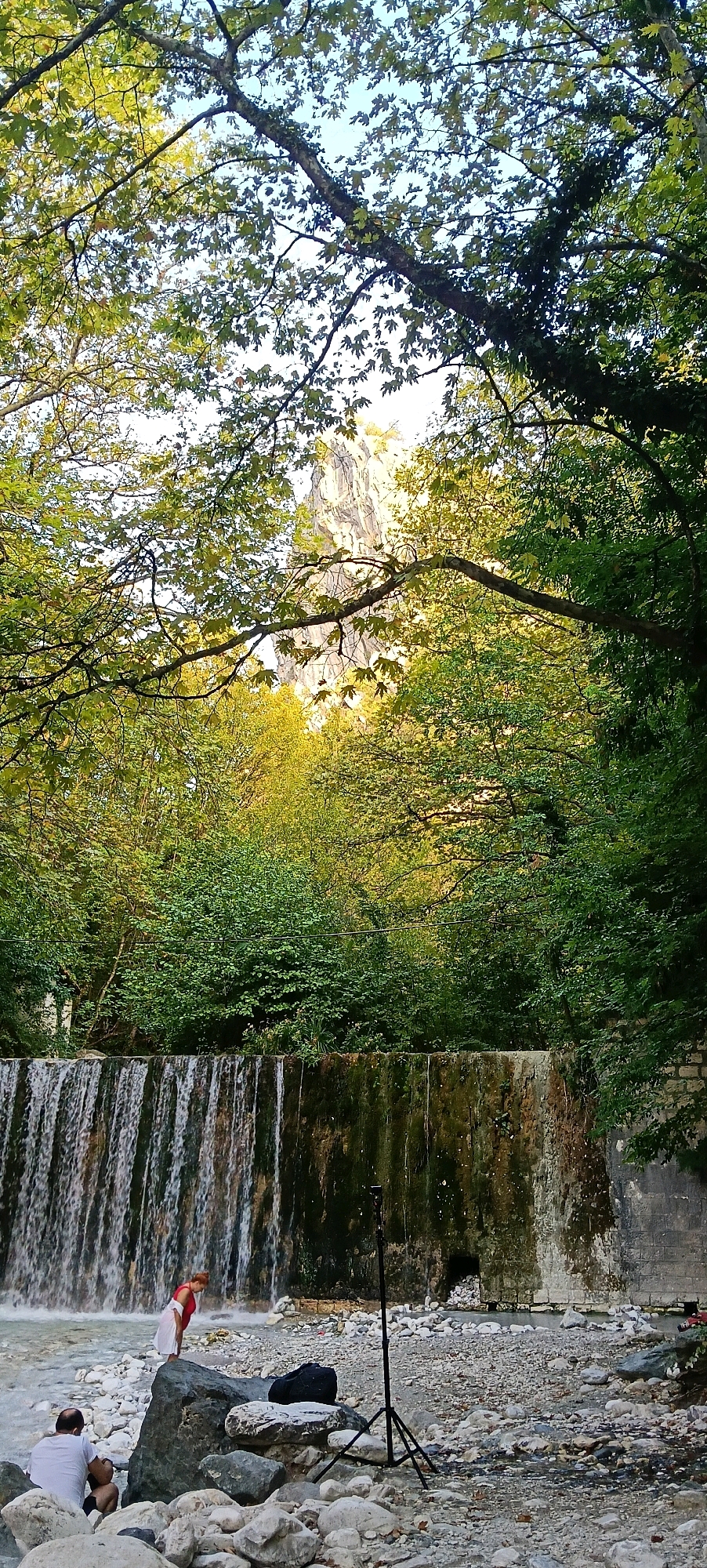 The image depicts a natural scene with a waterfall in the foreground and lush green trees and a rocky mountain in the background. The waterfall is made of a stone structure with water cascading down. Two people are present in the lower left corner of the image. One person is sitting on a rock, wearing a white shirt, while the other person, wearing a red top and a white skirt, stands near the waterfall. A tripod and a black bag are placed on the rocky ground near the waterfall. The environment is characterized by a river flowing in front of the waterfall, surrounded by rocks and vegetation. The presence of trees suggests a forest or park-like setting. The mountain in the background appears rugged and imposing. The overall atmosphere is serene and natural. It's likely the location is within Pozar Baths, Greece, given the natural setting with a waterfall.