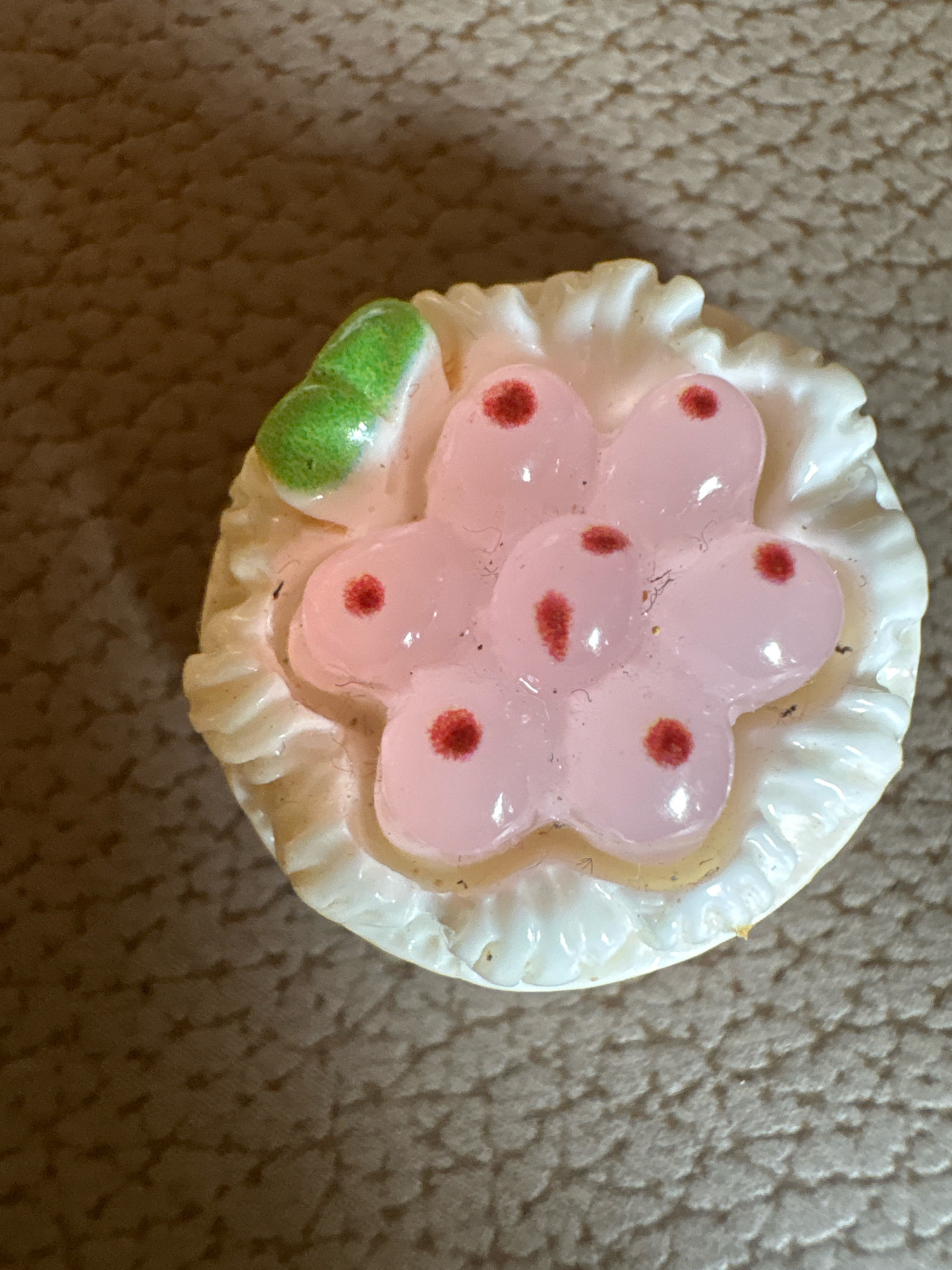 A macro photograph shows a miniature replica of a cupcake with a decorative pink icing and red dots arranged in a floral pattern. The cupcake is placed on a textured, brown surface that appears to be a piece of fabric or mat. A small green leaf adorns the top left of the cupcake. The lighting casts a subtle shadow to the left of the cupcake. The location of Vagator, India, is not discernible from the visual information provided in the image.
