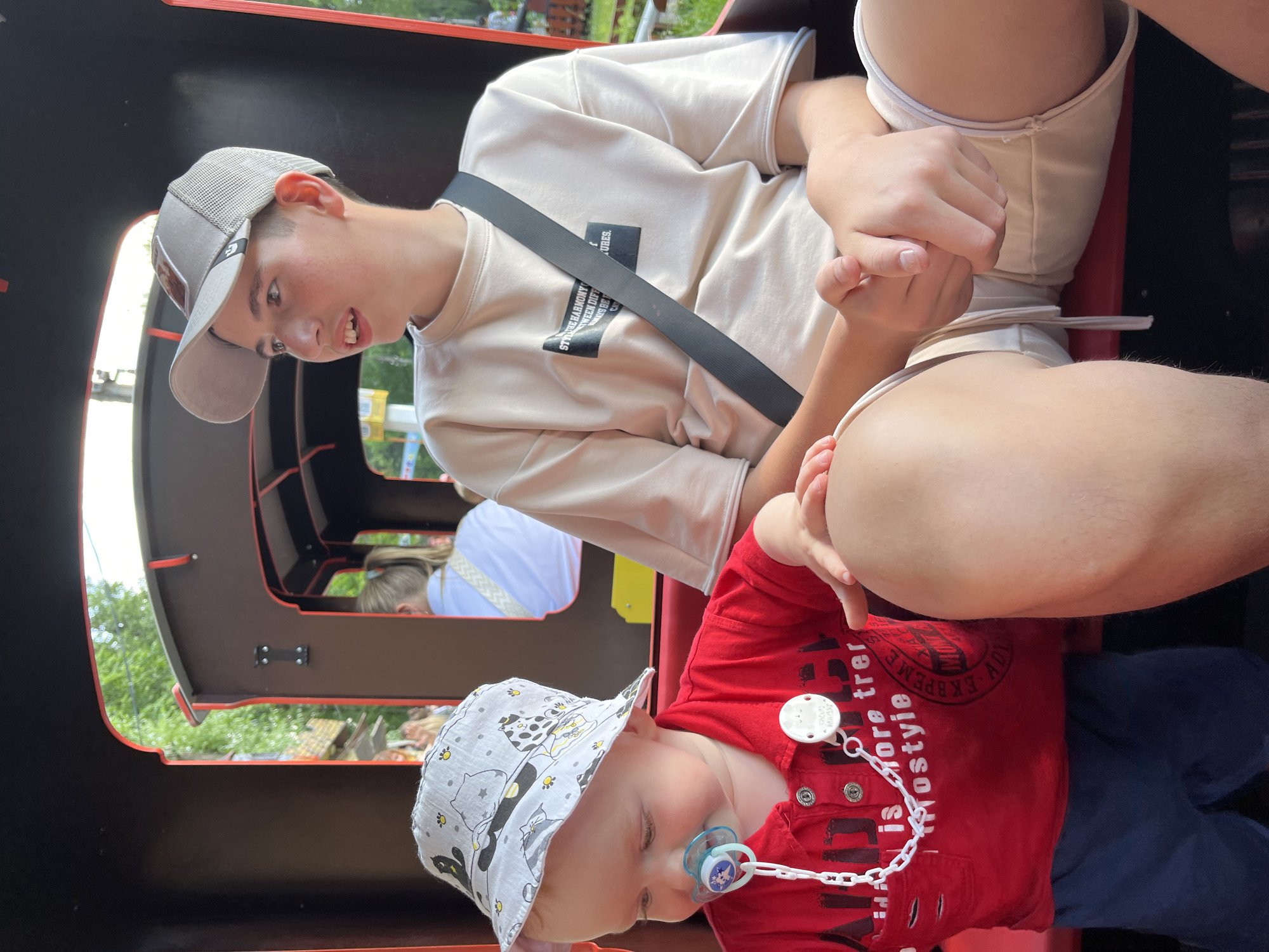 In a brightly lit outdoor scene, two children are seated in what appears to be a train car, possibly at an amusement park. The older child, wearing a grey baseball cap and a light-colored t-shirt with black text that reads "STYME HARMONY WEEN DIFF KYS BEF C URES.", leans forward, looking down with an amused expression. Their hands are intertwined with another person's, suggesting a close relationship. The younger child, in the foreground, wears a red shirt with similarly indistinct text, a white bucket hat adorned with a cat pattern, and a pacifier clipped to their clothing with a white chain. The baby's pacifier has a blue and white design. The background shows greenery, hinting at a natural or park-like setting. The presence of a train car and the overall atmosphere suggest a fun outing, perhaps a family trip. The lighting indicates it's daytime, likely sunny. The text on the clothing is difficult to decipher completely, but it contributes to the casual, youthful vibe of the image.