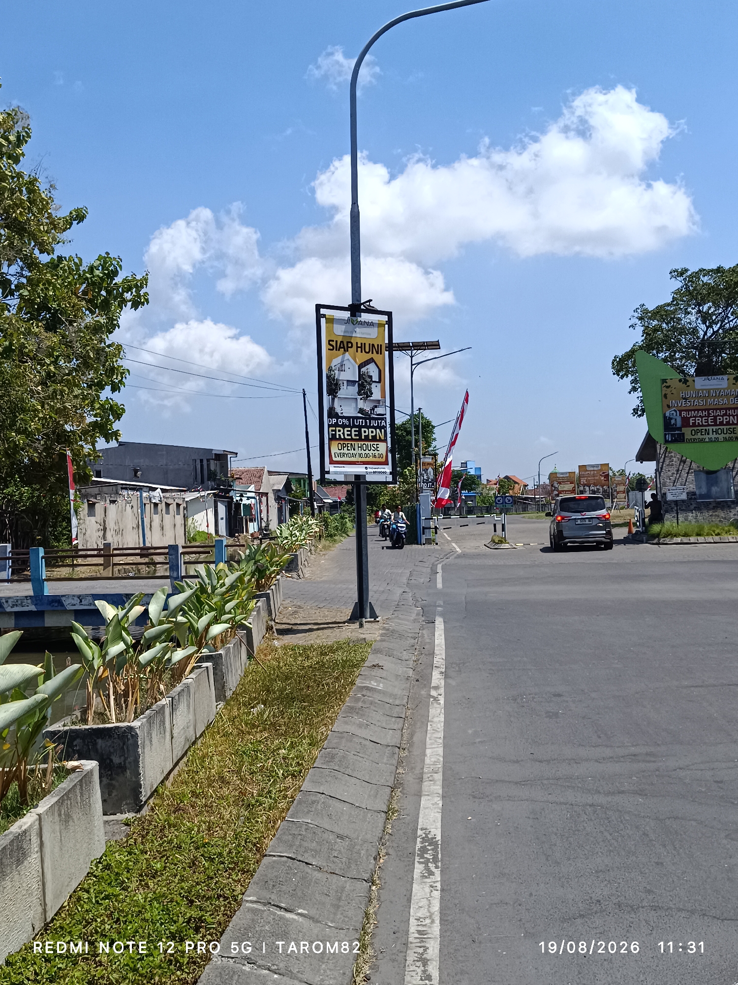 The image depicts a street scene in Tebel, Indonesia, on a bright, sunny day with scattered clouds. A prominent street lamp with a banner advertising "JIVANA SIAP HUNI" (JIVANA Ready to Occupy) is positioned on the right side of the road. The banner details an "OPEN HOUSE" with specific operating hours and mentions "DP 0%" and "FREE PPn". To the left of the street lamp, a similar banner is visible.
The road is paved and a white line marks the edge. On the left side of the road, a concrete planter box filled with green foliage runs along the sidewalk. Further left, a blue and white striped barrier is visible, and behind it, several low-rise buildings with varying roof styles and some showing signs of disrepair. A red and white Indonesian flag is planted on a pole near the barrier.
In the middle ground, a silver car is driving away from the camera. Two motorcycles are also on the road, one with a rider and passenger. A gated entrance with a barrier arm is present further down the road, and a person appears to be standing near the entrance.
The environment suggests a residential or commercial development area, possibly an estate or complex, given the advertising banners and the gated entrance. The overall impression is one of an active street with some ongoing development or sales activity. The date stamp on the image indicates August 19, 2026, at 11:31 AM.