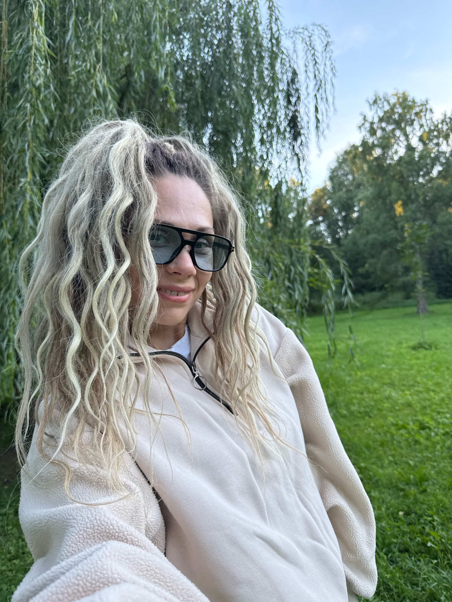 A woman with blonde, wavy hair is taking a selfie in an outdoor setting. She is wearing large, dark-framed sunglasses and a light-colored hooded jacket with a zipper. She is smiling, with her teeth visible. The background consists of lush green grass and trees, with a clear sky overhead. The image appears to have been taken in Rusanovo, Russia.