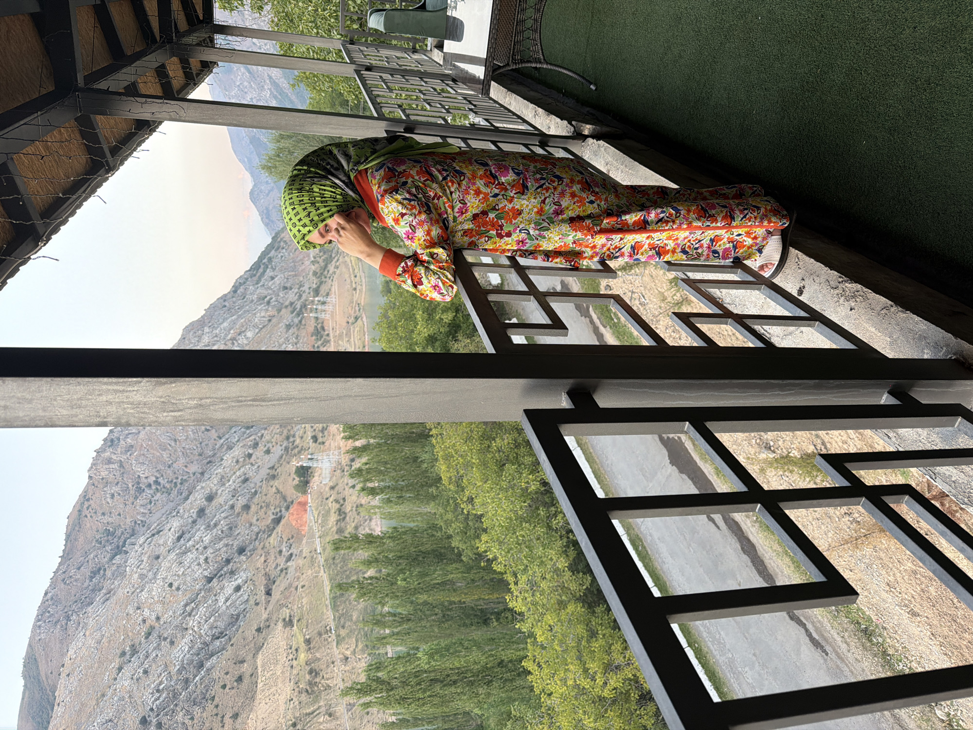 In Nanay, Uzbekistan, a woman stands on a balcony, gazing at a mountain landscape. She is wearing a vibrant, floral-patterned dress and a green headscarf. The balcony features a metal railing and a dark-colored roof with decorative lights. Below the balcony, a road winds through a valley dotted with tall, slender trees and lush green foliage. In the background, rugged, rocky mountains rise under a pale sky. A body of water, possibly a lake or river, is visible in the distance, reflecting the muted tones of the sky. The scene conveys a sense of tranquility and appreciation for the natural beauty of the region.