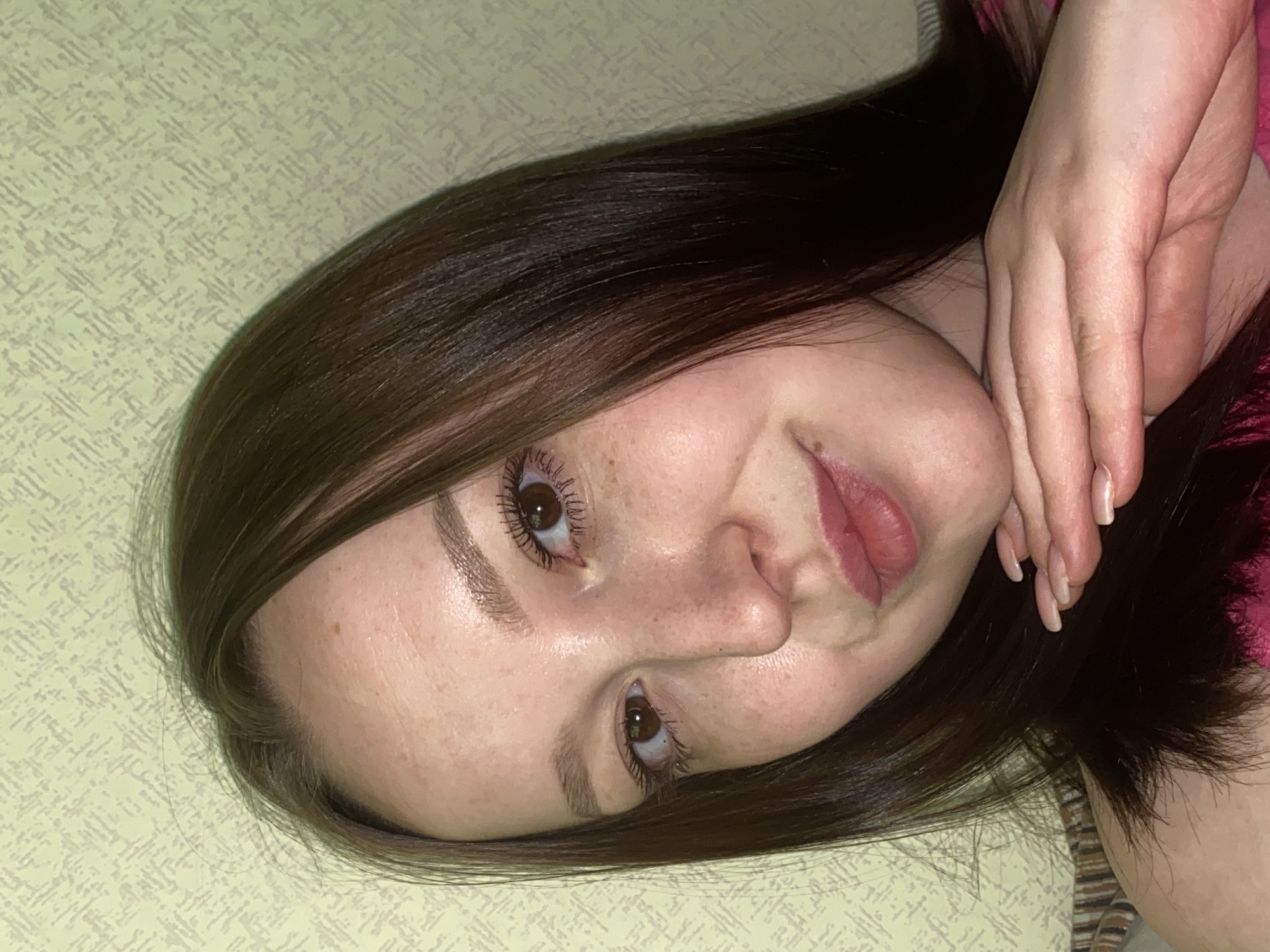 A young woman with brown hair and freckled skin is lying down, angled towards the camera. She has brown eyes and is wearing mascara. Her lips are slightly parted, and she is resting her hand near her face. The background is a light-colored patterned surface. The location in Chaikovsky, Russia, cannot be determined from the visual information provided.
