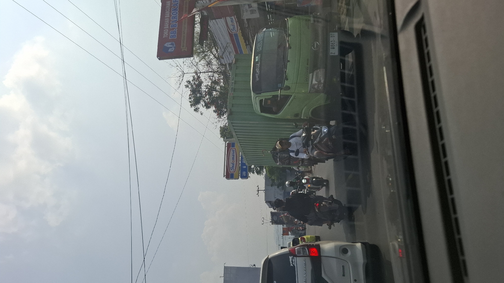 The image captures a busy street scene in Cikaroya, Indonesia, on a bright, sunny day with scattered clouds. The perspective is from inside a vehicle, looking forward. The foreground is dominated by the dashboard of the car, with its ventilation grilles visible.
The middle ground of the image is filled with traffic. Several motorcycles are prominent, with riders dressed in casual clothing and helmets. One motorcycle in the right middle ground has a rider wearing a beige cap and a white shirt, carrying a backpack. Another motorcycle further to the left shows a rider in a black jacket. In the center of the scene, a large, light green truck with a container trailer is moving. The truck has Arabic text on its windshield. Its license plate is visible as "Z 8861 DT".
On the right side of the road, a red and white sign for "TOKO BAHAN BANGUNAN BB. BAROKAH" (Construction Materials Store BB. Barokah) is clearly visible, along with a smaller red and white "Indomaret" sign below it. There are also other signs and buildings lining the street. On the left side of the image, the rear of a white vehicle, likely an SUV, is partially visible.
The environment suggests a commercial or mixed-use area within the city, with overhead power lines crisscrossing the sky. The overall impression is one of moderate traffic congestion on a typical day.