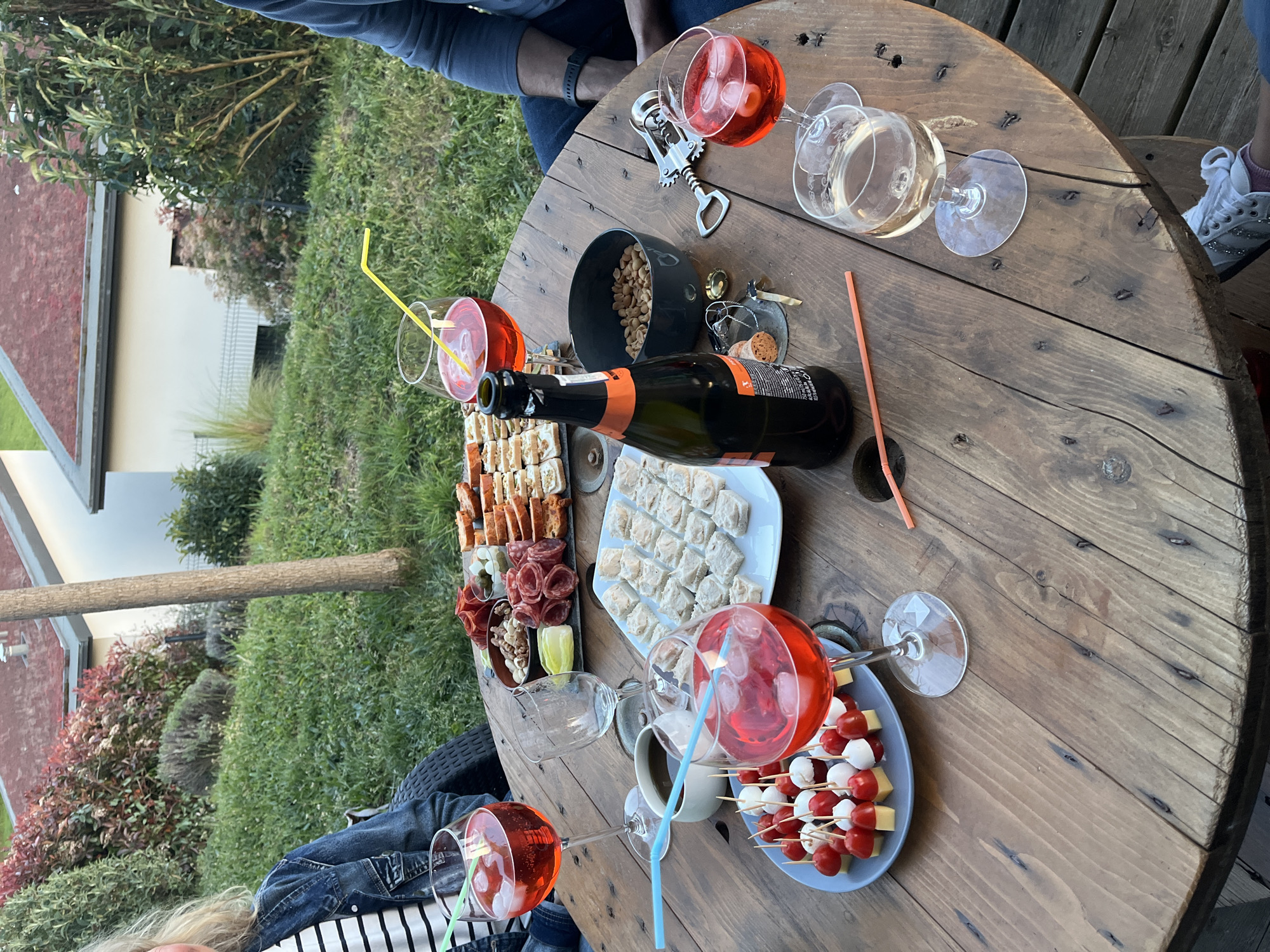 The image depicts a social gathering centered around a round, wooden table laden with food and drinks. Located outdoors in Chambœuf, France, the table is surrounded by people, partially visible.
On the table, there are several glasses filled with a red beverage, likely spritzers, as well as glasses of white wine. A bottle of sparkling wine rests among plates of snacks, including skewers of tomatoes and mozzarella, assorted appetizers, and a bowl of nuts. Utensils like a bottle opener and cork are also present.
The environment consists of an outdoor space with greenery, parts of a house visible in the background, and elements that suggest a relaxed, informal setting.