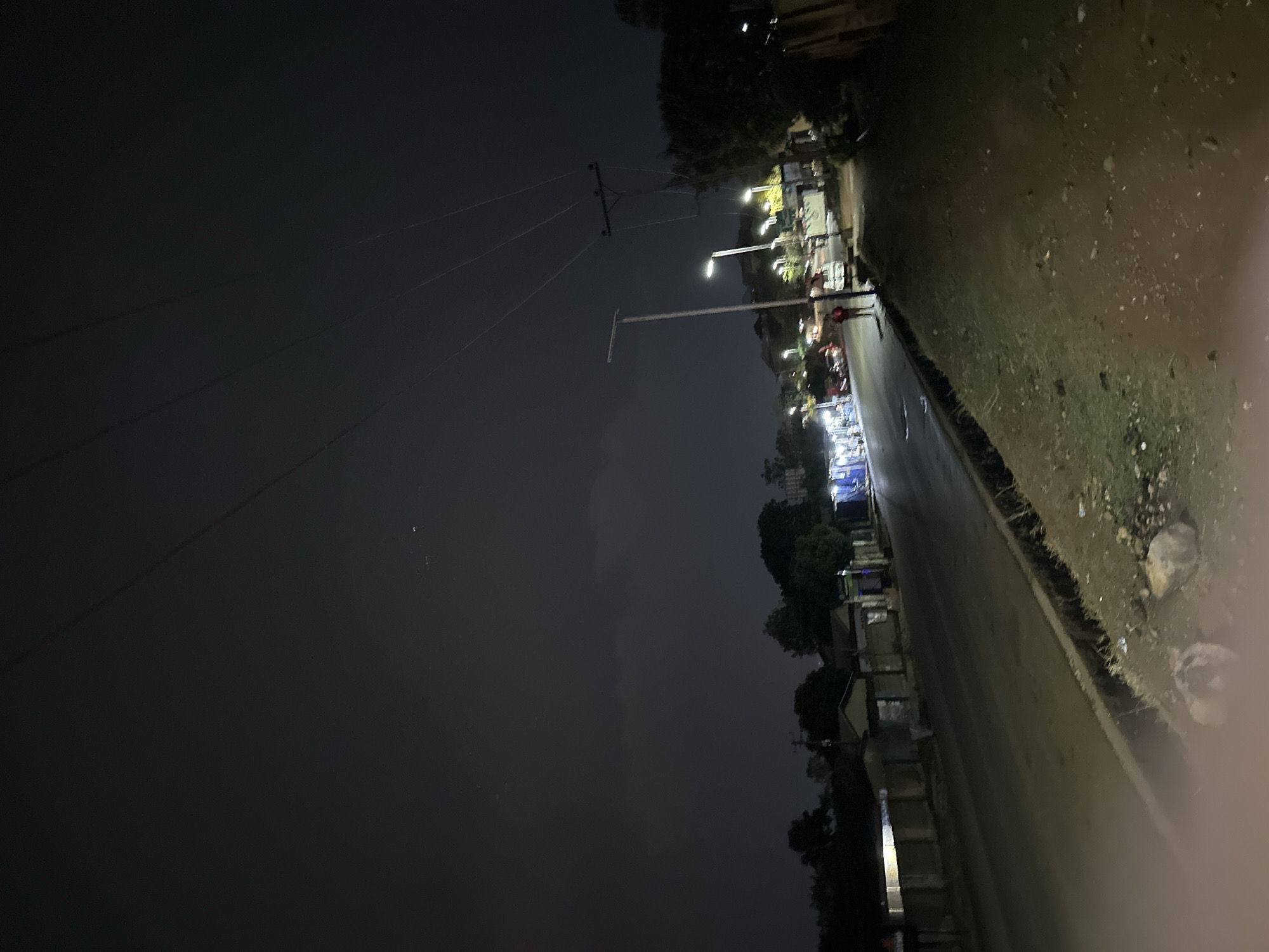 This is a nighttime view in Jos, Nigeria. The scene captures a street lined with buildings. The sky is dark, with utility poles and power lines visible. Streetlights illuminate the area. The road is paved and there is vegetation. There are also buildings lining the streets with some illuminated.