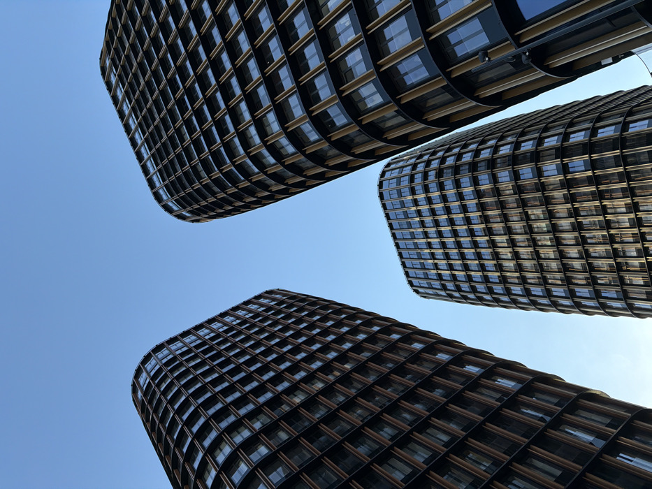 Three modern, curved high-rise buildings are depicted from a low-angle perspective against a clear blue sky. These structures are the Capital Towers, located in the Presnensky District of Moscow, Russia. Each tower features a dark, multi-story facade composed of numerous rectangular windows and prominent vertical golden-hued architectural fins. Two larger towers partially frame the view on the left and right, with a third, slightly shorter tower visible between and behind them in the center. No individuals, vehicles, or ground-level urban elements are present within the frame.