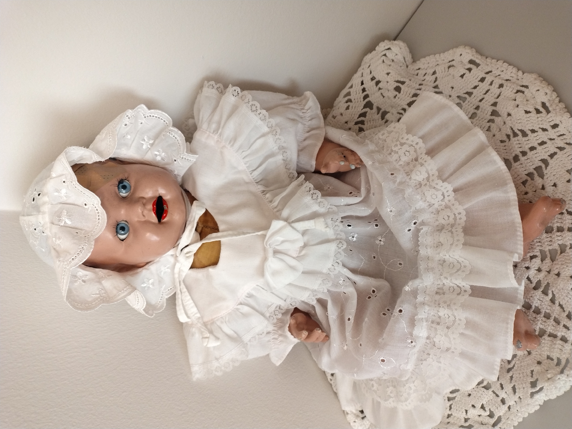 A vintage doll with blue eyes and red lips, dressed in a white lace-trimmed christening gown and bonnet, is seated on a white crocheted doily. The doll is positioned in the upper half of the frame, against a plain white background. The doll's face is slightly aged, with visible paint wear on the lips and skin. Its hands are visible, resting on its lap. The lighting suggests an indoor setting, possibly a display or storage area. The image does not contain any specific or identifiable landmarks, buildings, or street scenes that could be used to ascertain its location within Písek, Czechia.