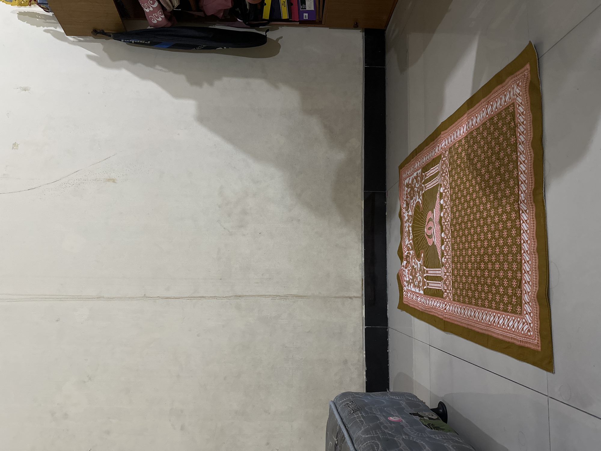 The image depicts an indoor scene, likely a room in a residence in Bulakrejo, Indonesia. In the foreground, a prayer mat with a brown and pink floral pattern is laid out on a tiled floor. To the left, a portion of a gray upholstered sofa or bed is visible. The background features a plain, off-white wall with some subtle staining and a vertical water streak. To the right, a wooden cabinet or shelf unit is present, with a dark bag hanging from it and several items, including boxes and folded fabric, stored on its shelves. The lighting creates a shadow on the wall, suggesting a light source from the right.