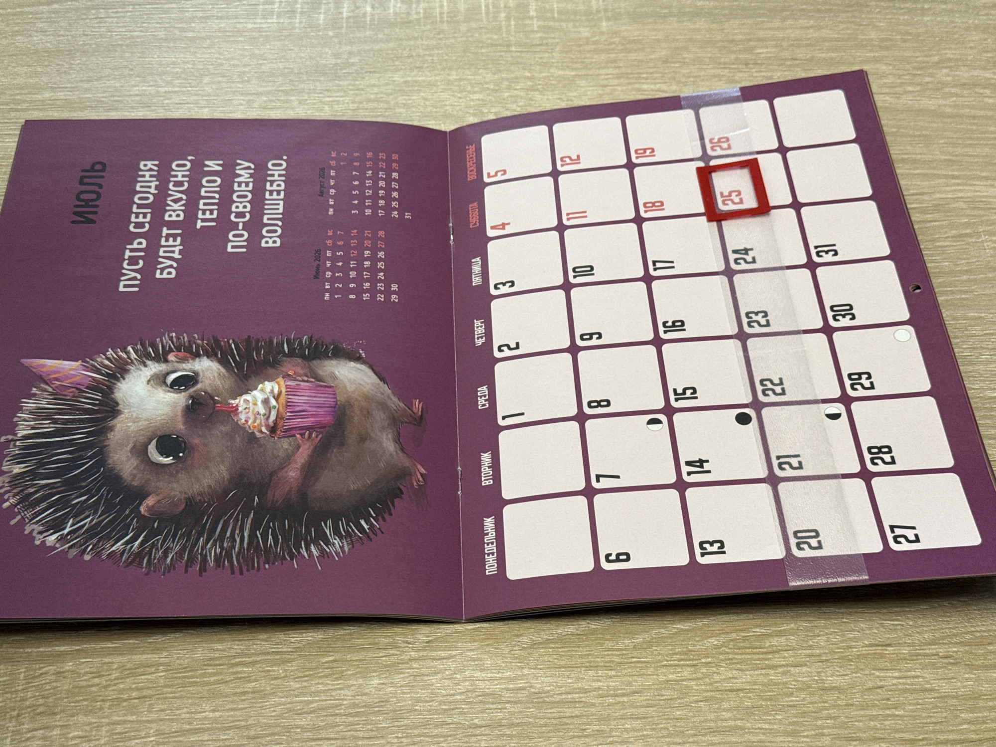 The image shows a wall calendar page for July. The top portion of the page features an illustration of a hedgehog holding a cupcake and text in Russian that reads "Пусть сегодня будет вкусно, тепло и по-своему волшебно" which translates to "May today be tasty, warm and magical in its own way." Below the illustration is a calendar grid showing the days of the week from Monday to Sunday. The month is July, as indicated by the title "ИЮЛЬ" at the top right of the page. The date '25' is highlighted with a red square. The calendar is attached to a wooden surface, likely a wall. There are no people visible in the image. The overall scene depicts a calendar page from a dwelling or office. The image does not contain any information that allows for determining its location within Lyubertsy, Russia.