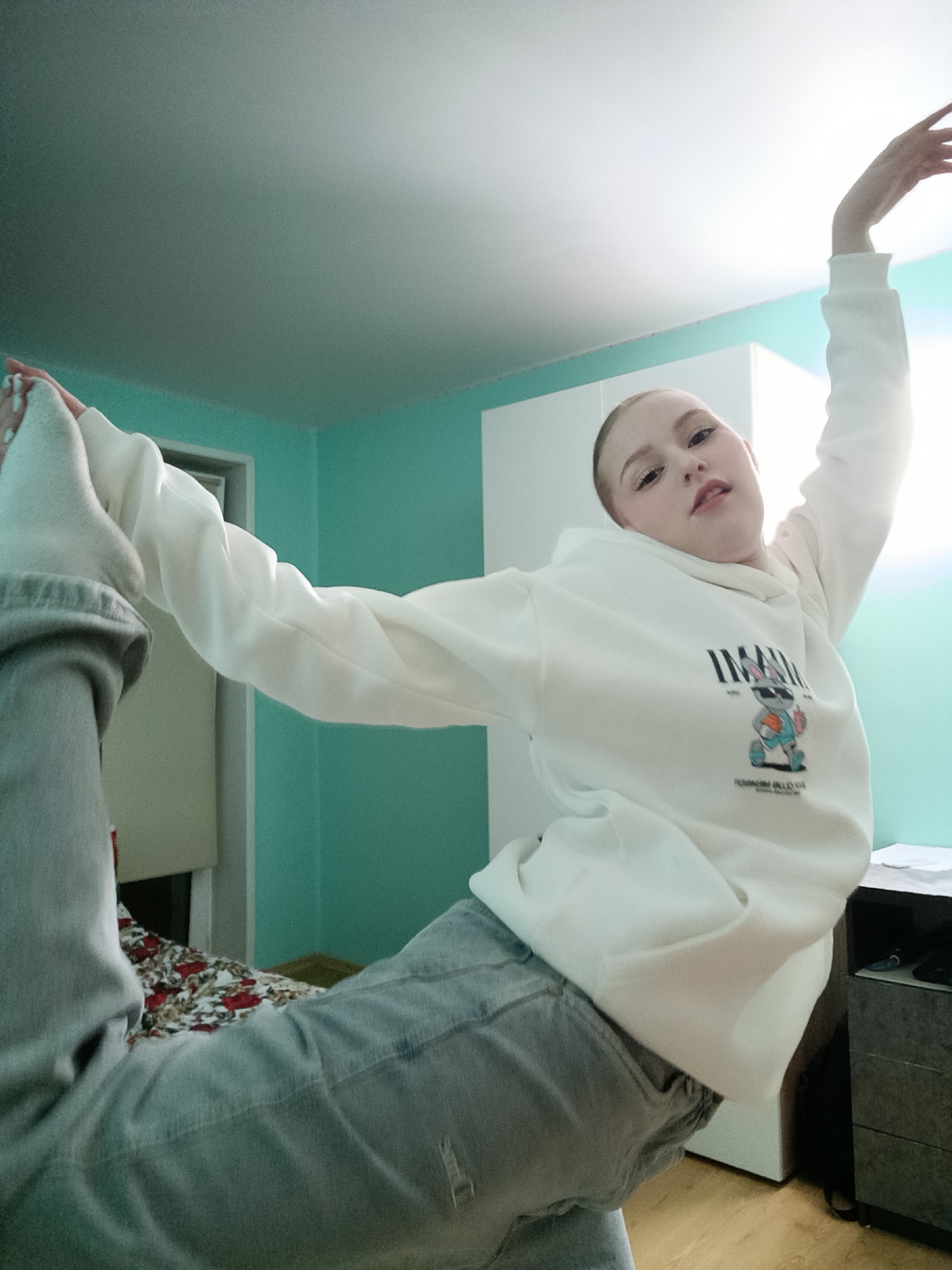 A person is indoors, performing a flexible pose. They are wearing a white hooded sweatshirt with a graphic design on the front, light gray jeans, and white socks. One leg is extended upwards, held by a hand, while the other leg is bent. The person's other arm is raised upwards. The room has teal-colored walls and light wooden floors. In the background, there is a white wardrobe, a nightstand, and a bed with a floral patterned bedspread. Based on the available information, it's not possible to independently verify the location as Chaikovsky, Russia.